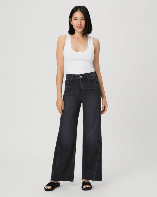 A woman modeling a pair of dark charcoal wide-leg jeans with a frayed hem, paired with a white tank top and black slide sandals, against a light gray studio background.