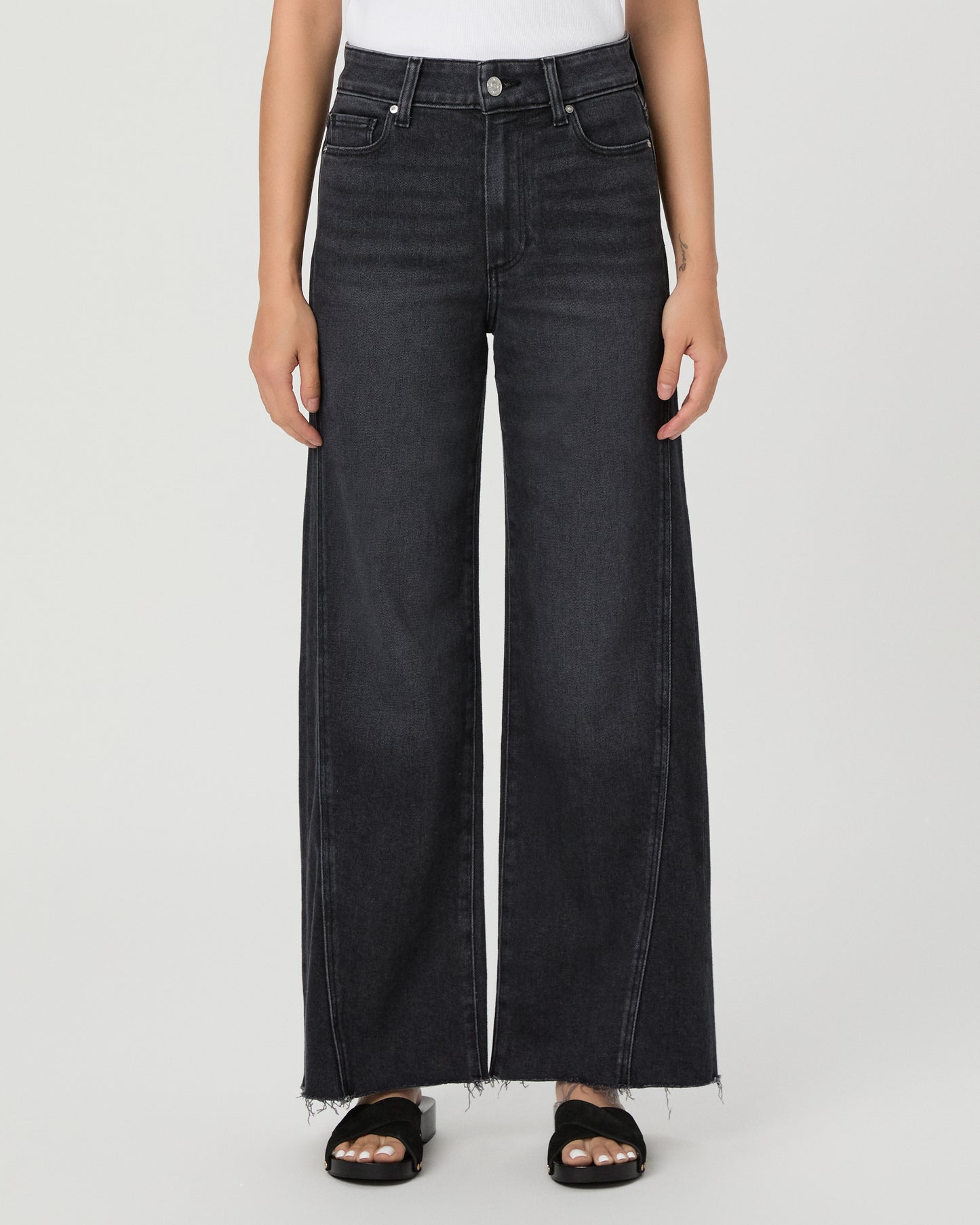 A woman modeling a pair of dark charcoal wide-leg jeans with a frayed hem, paired with a white tank top and black slide sandals, against a light gray studio background.