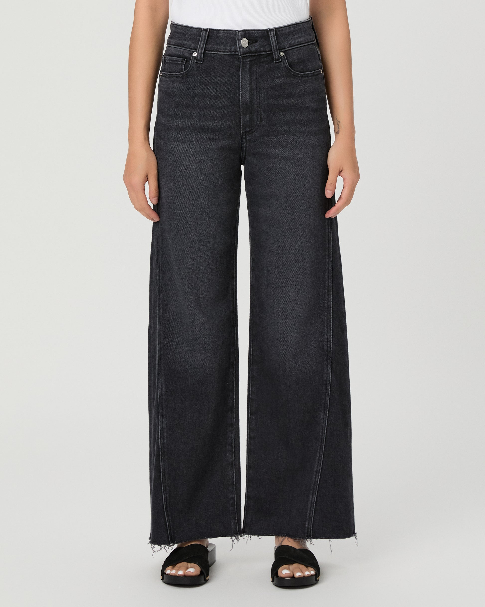 A woman modeling a pair of dark charcoal wide-leg jeans with a frayed hem, paired with a white tank top and black slide sandals, against a light gray studio background.