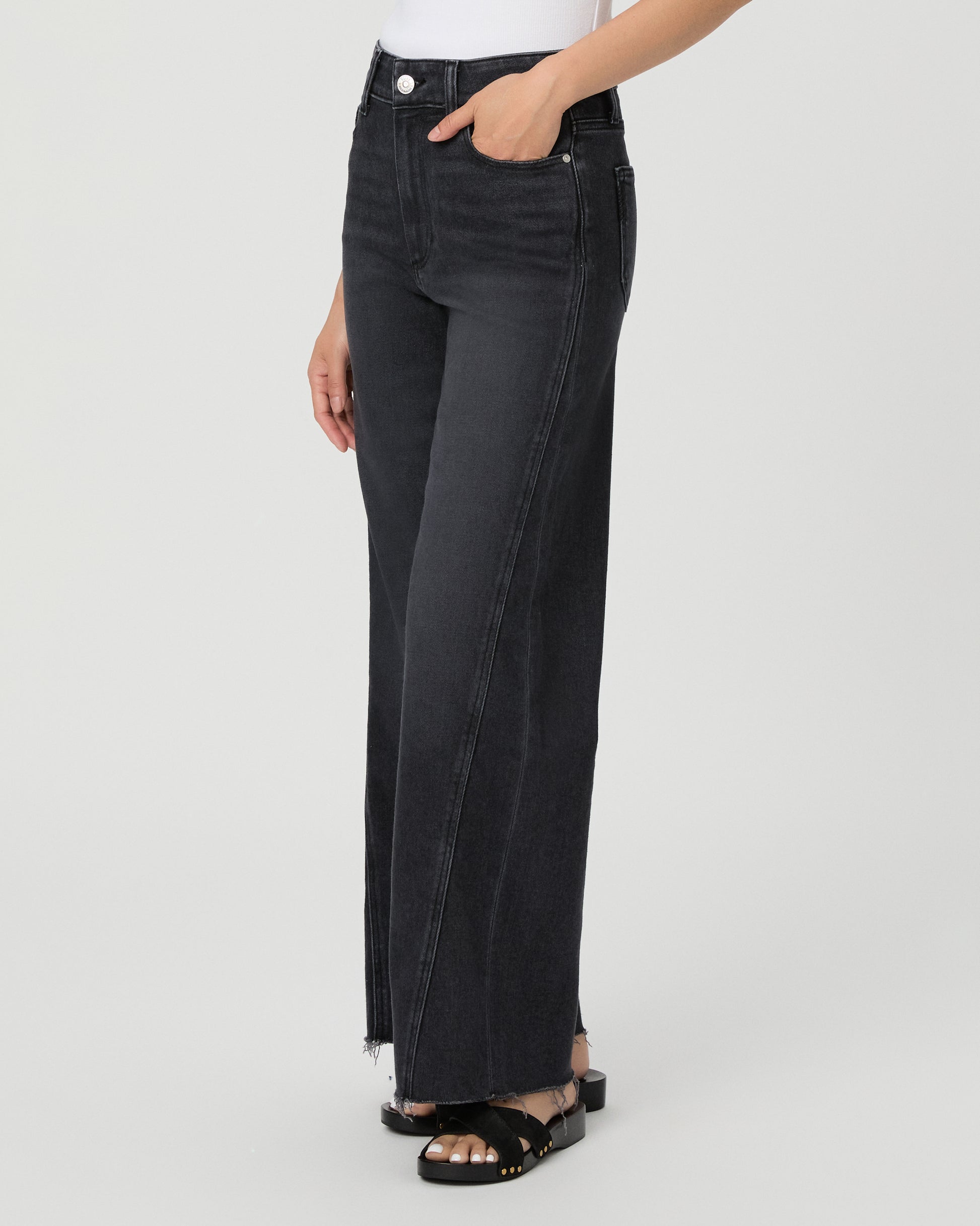 A woman modeling a pair of dark charcoal wide-leg jeans with a frayed hem, paired with a white tank top and black slide sandals, against a light gray studio background.