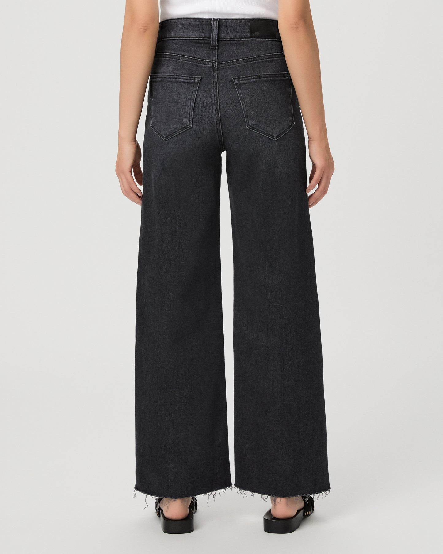 A woman modeling a pair of dark charcoal wide-leg jeans with a frayed hem, paired with a white tank top and black slide sandals, against a light gray studio background.