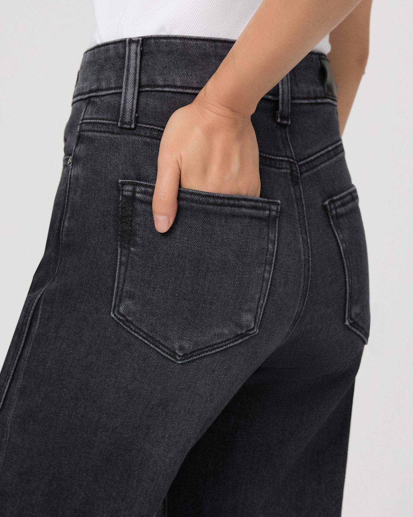A woman modeling a pair of dark charcoal wide-leg jeans with a frayed hem, paired with a white tank top and black slide sandals, against a light gray studio background.