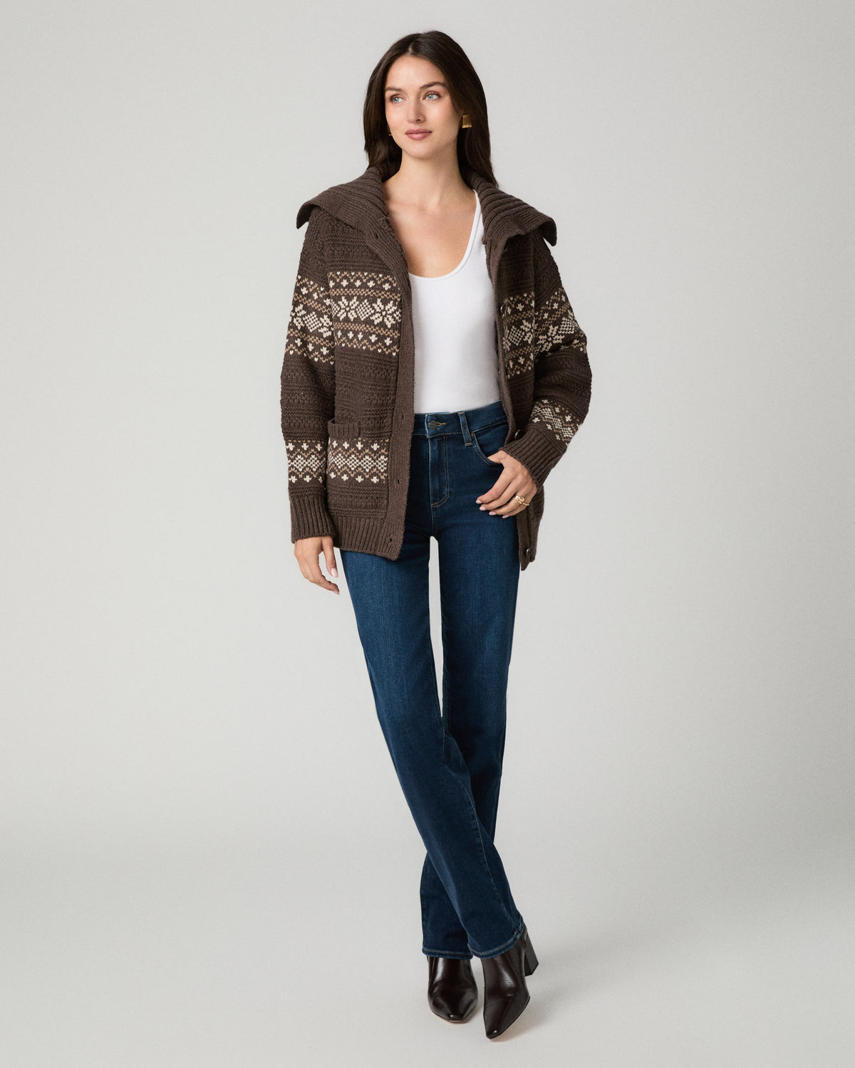 A woman modeling a brown knit zip-up cardigan with a white tank top, blue jeans, and dark brown pointed-toe heels against a light gray studio background.