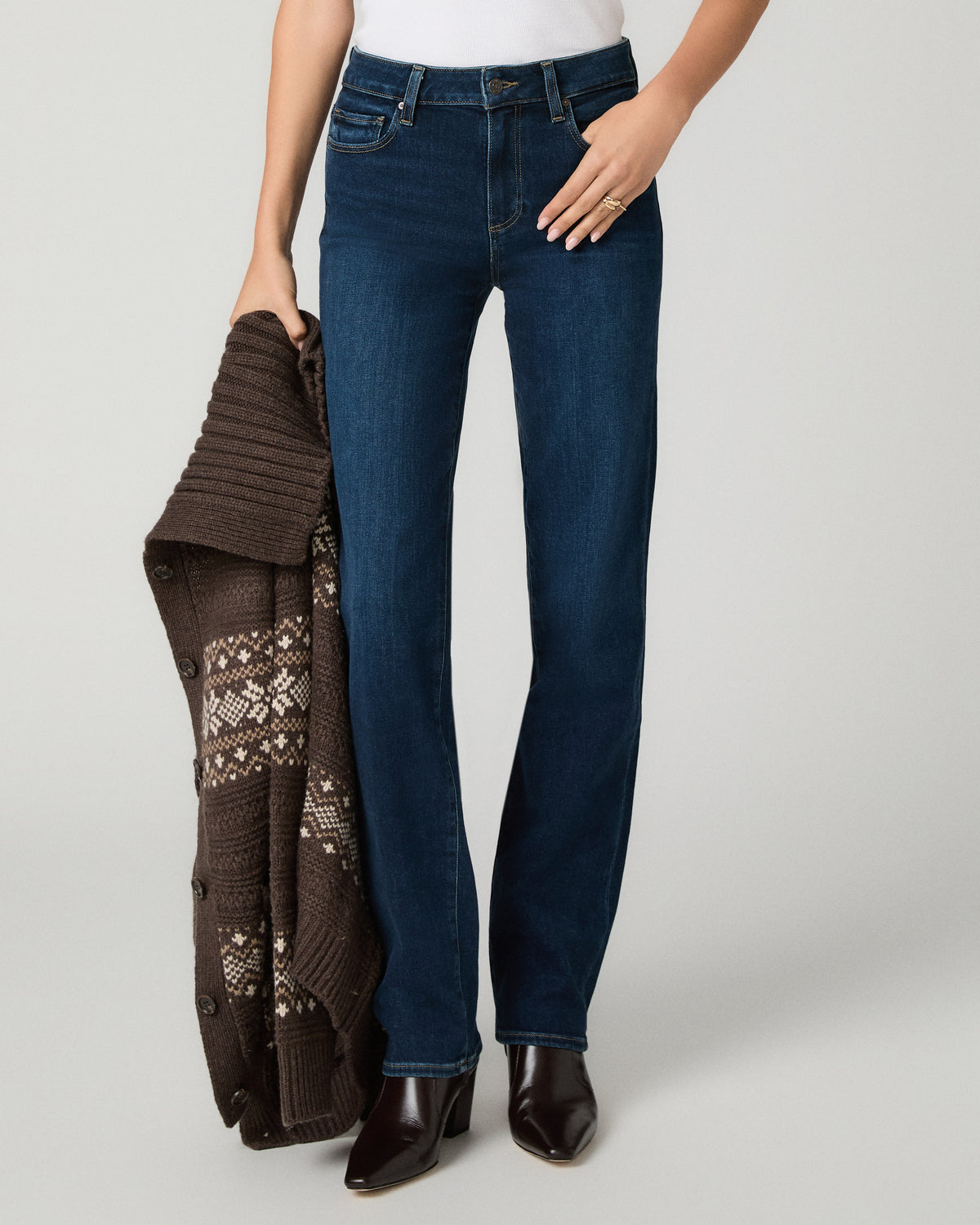 A model wearing dark blue straight-leg jeans, holding a brown knit cardigan with a geometric pattern, standing against a light gray studio background.
