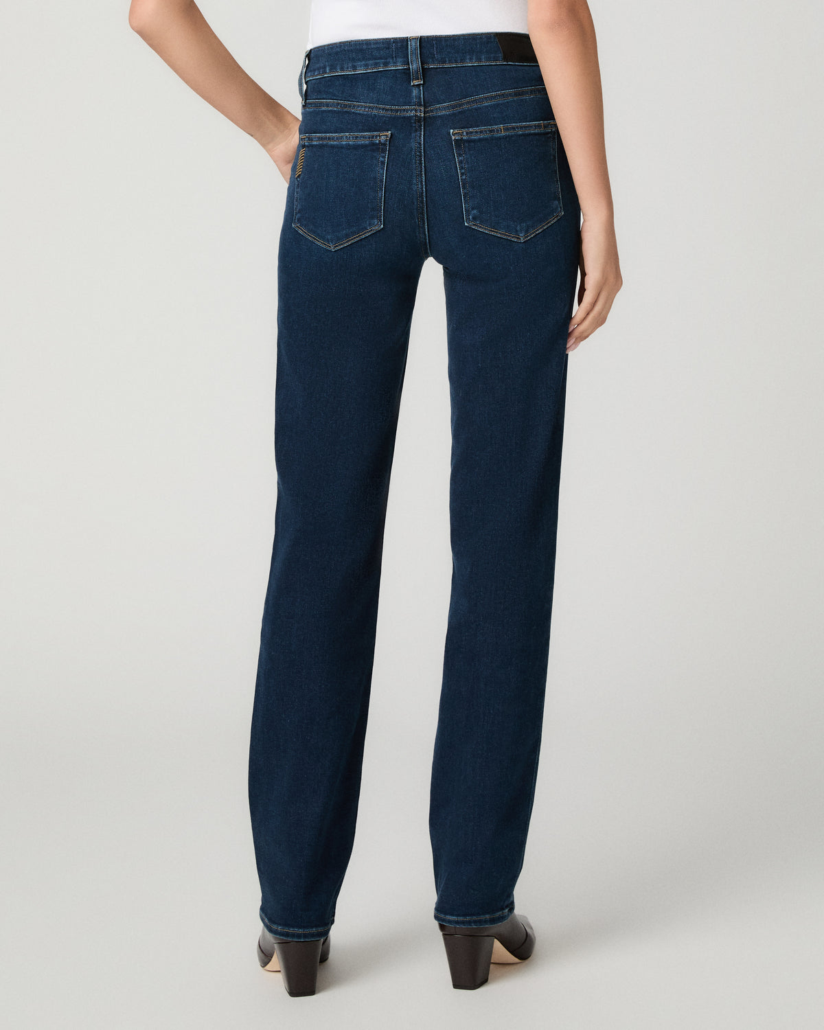 Rear view of a model wearing dark blue straight-leg denim jeans with a white top and black heels, photographed against a light gray studio background.