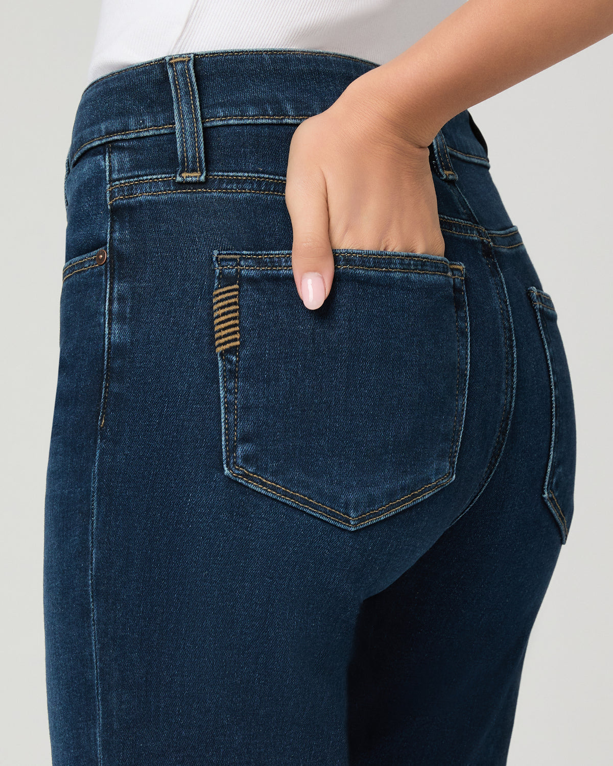 Close-up back view of a person wearing dark blue denim jeans, showing the back pocket with a hand inserted and a small striped tab detail on the pocket.