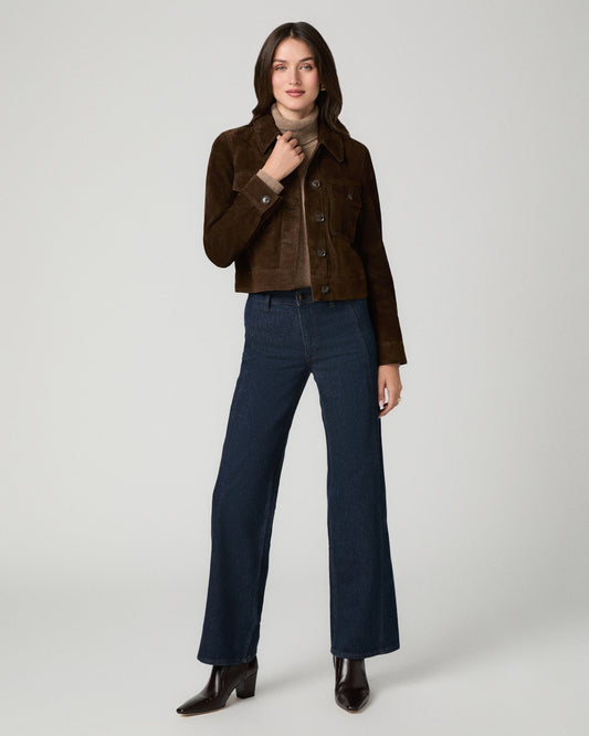 A model stands against a light gray studio background wearing a cropped brown suede jacket over a beige turtleneck, dark blue wide-leg jeans, and dark brown ankle boots.