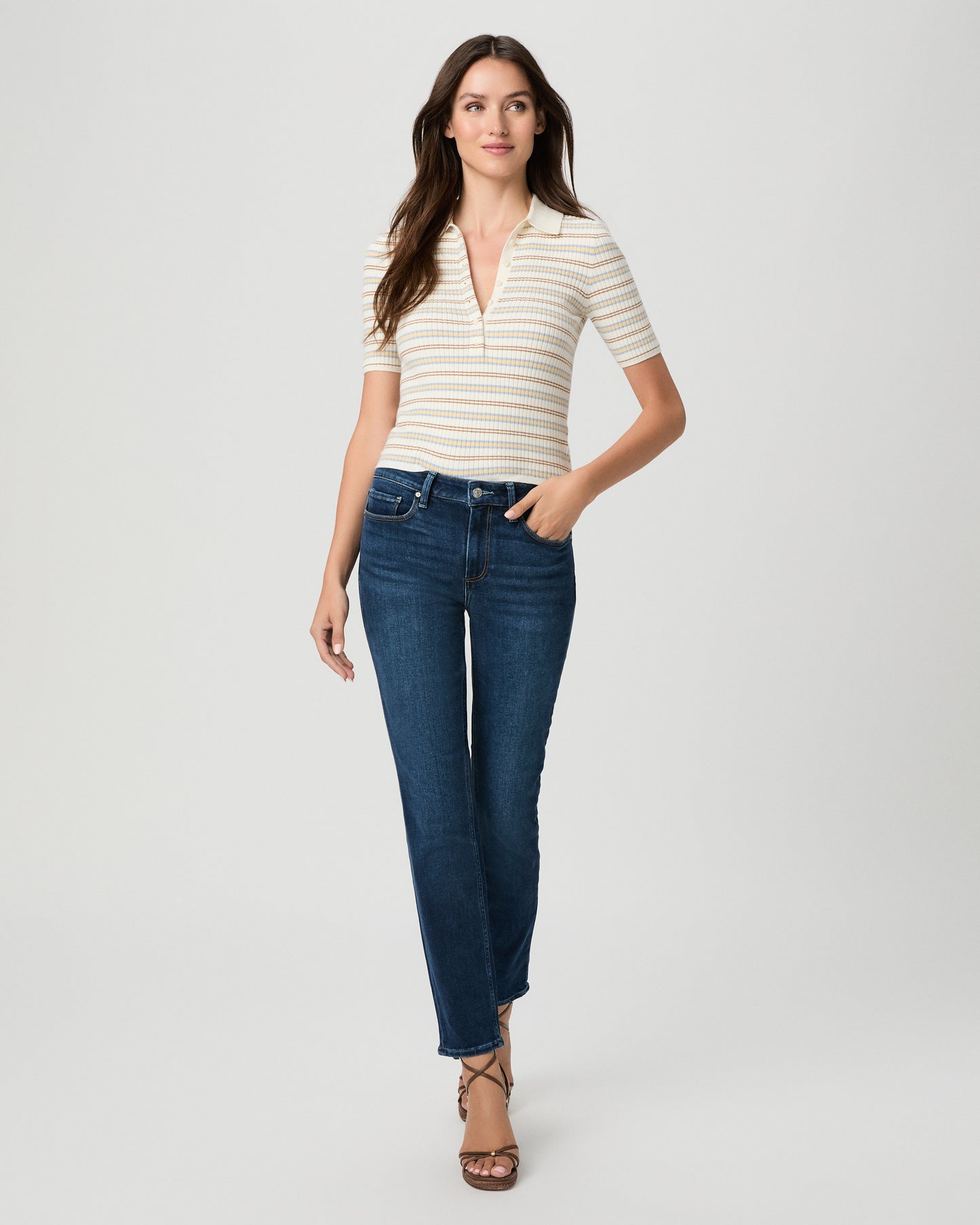 A woman modeling a short-sleeve polo shirt with thin cream and tan horizontal stripes, paired with dark blue straight-leg jeans and brown strappy heels, against a light gray studio background.