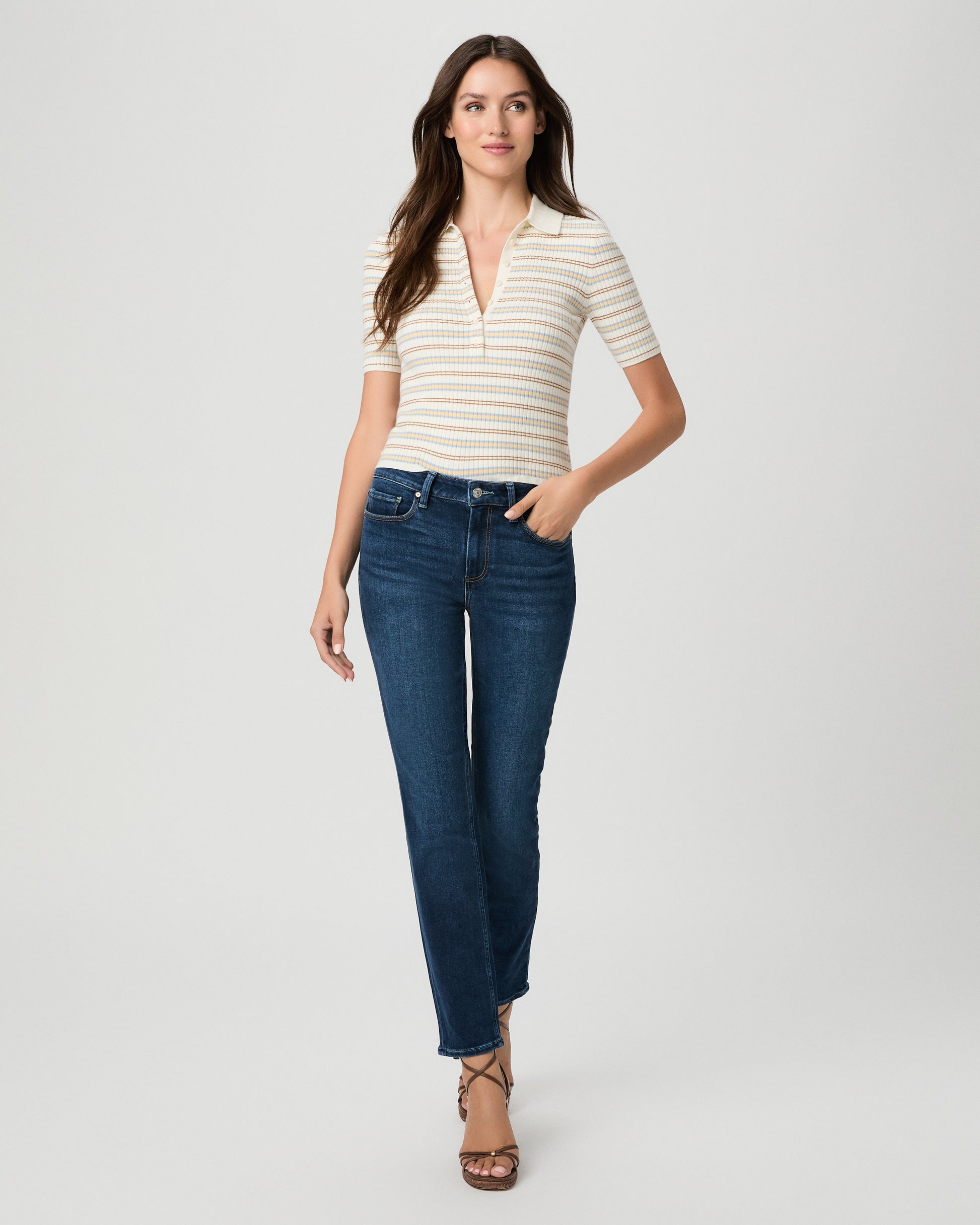 A woman modeling a short-sleeve polo shirt with thin cream and tan horizontal stripes, paired with dark blue straight-leg jeans and brown strappy heels, against a light gray studio background.