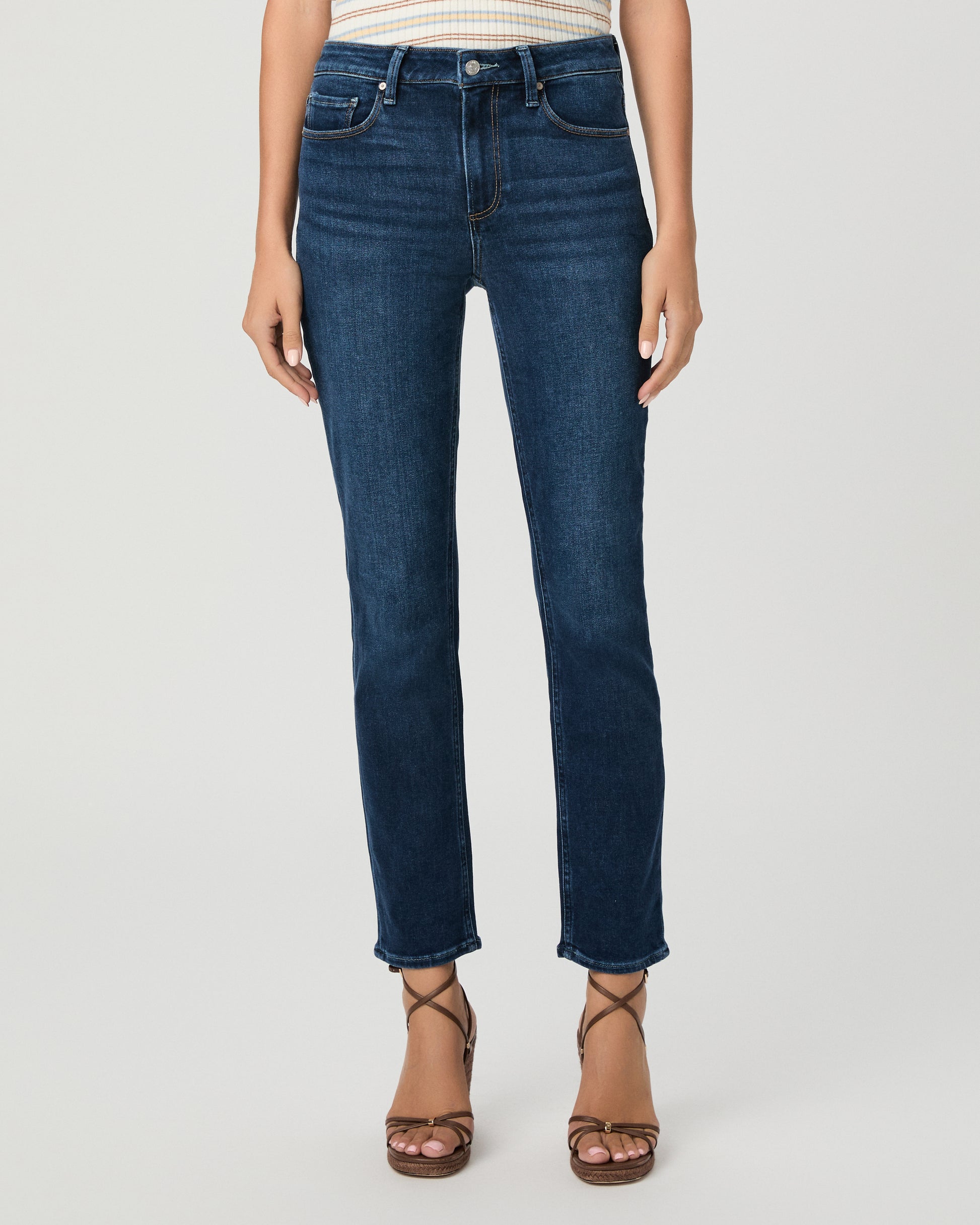 A woman modeling a short-sleeve polo shirt with thin cream and tan horizontal stripes, paired with dark blue straight-leg jeans and brown strappy heels, against a light gray studio background.