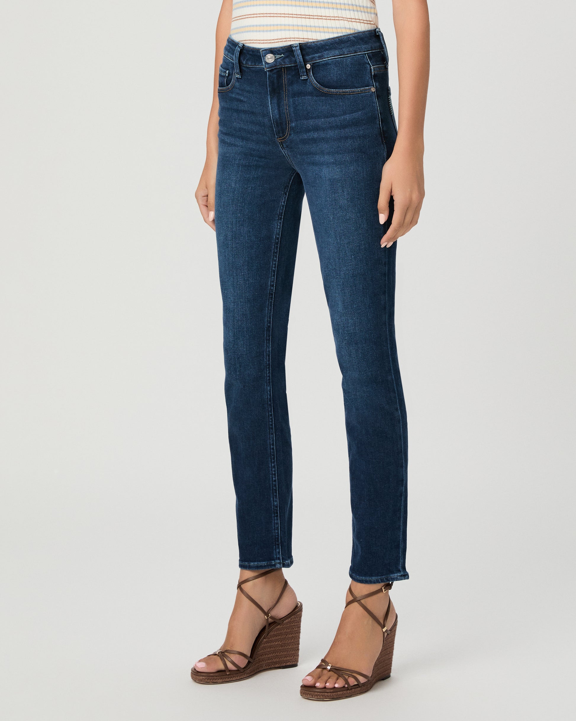 A woman modeling a short-sleeve polo shirt with thin cream and tan horizontal stripes, paired with dark blue straight-leg jeans and brown strappy heels, against a light gray studio background.
