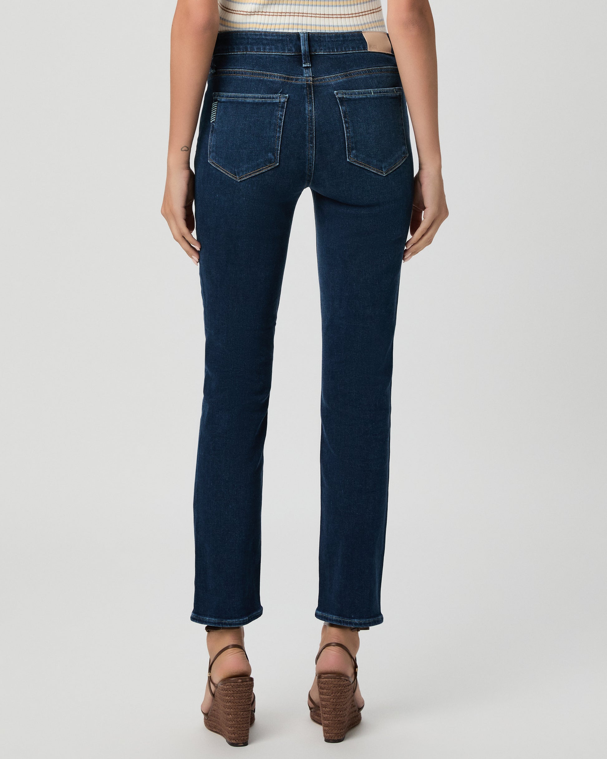 A woman modeling a short-sleeve polo shirt with thin cream and tan horizontal stripes, paired with dark blue straight-leg jeans and brown strappy heels, against a light gray studio background.