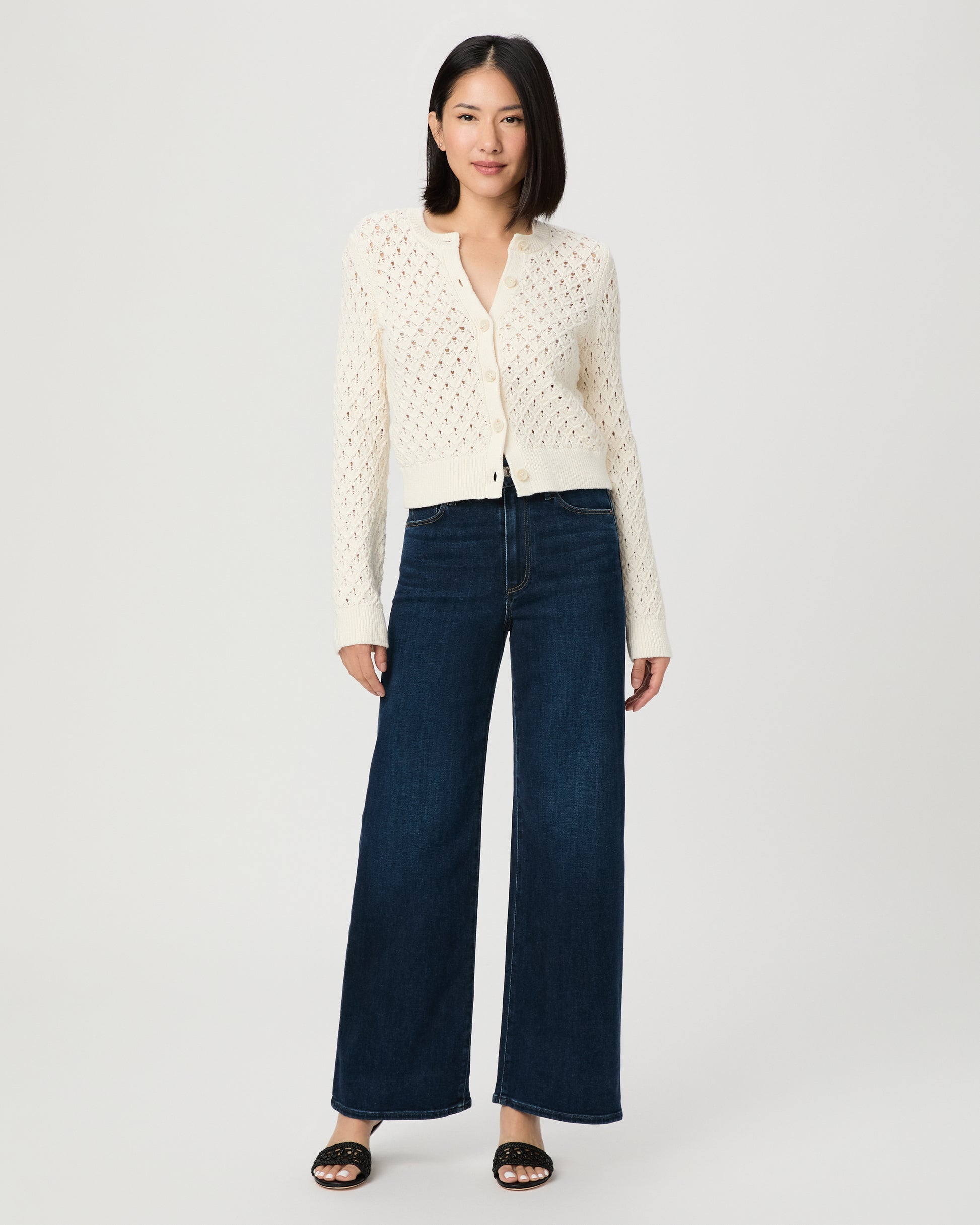 A woman modeling a cream-colored, long-sleeve open-knit cardigan paired with dark blue wide-leg jeans and black slide sandals, set against a plain light gray studio background.