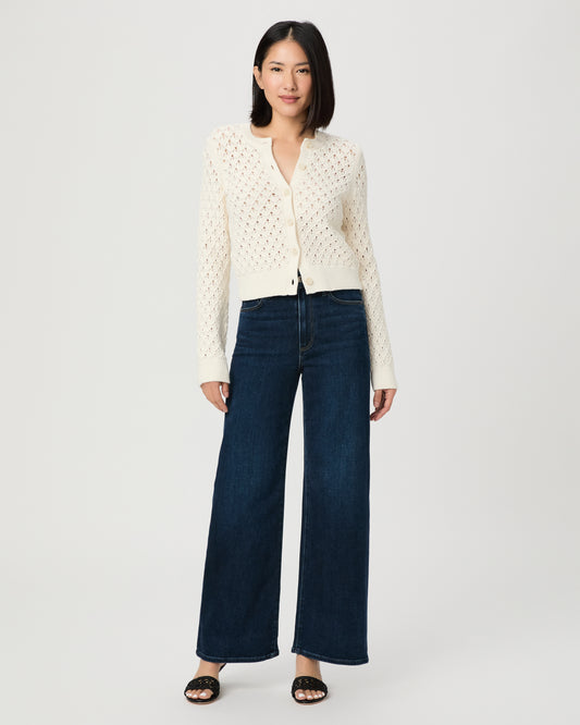 A woman modeling a cream-colored, long-sleeve open-knit cardigan paired with dark blue wide-leg jeans and black slide sandals, set against a plain light gray studio background.