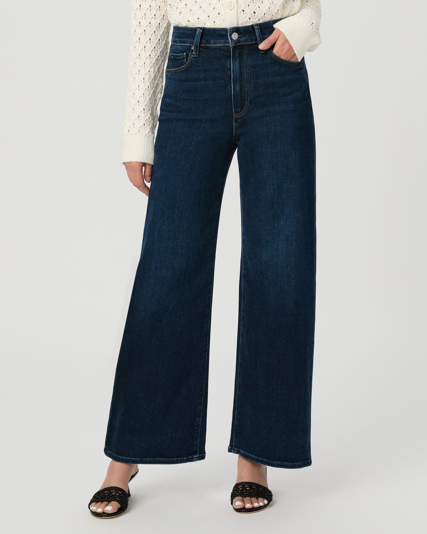 A woman modeling a cream-colored, long-sleeve open-knit cardigan paired with dark blue wide-leg jeans and black slide sandals, set against a plain light gray studio background.