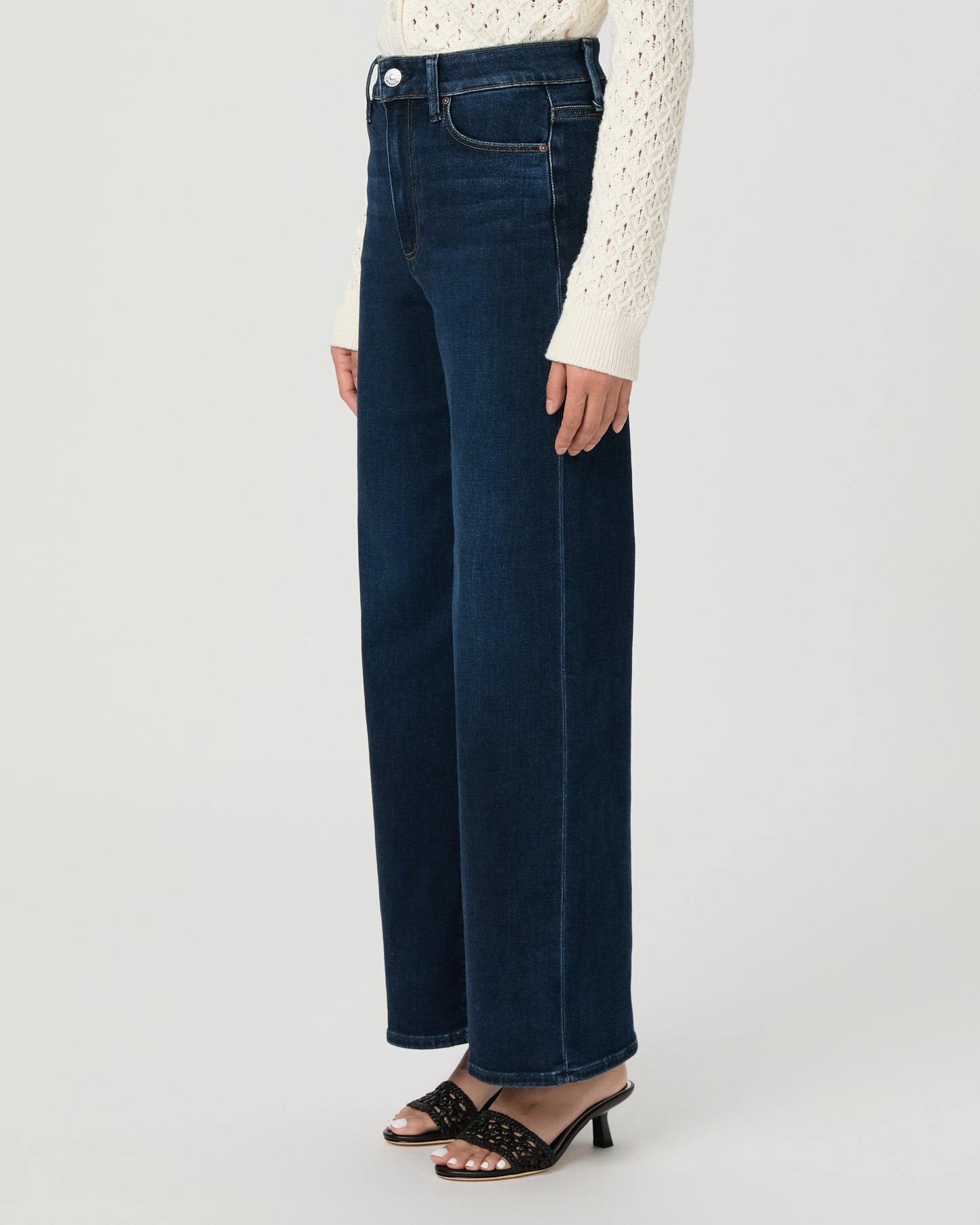 A woman modeling a cream-colored, long-sleeve open-knit cardigan paired with dark blue wide-leg jeans and black slide sandals, set against a plain light gray studio background.
