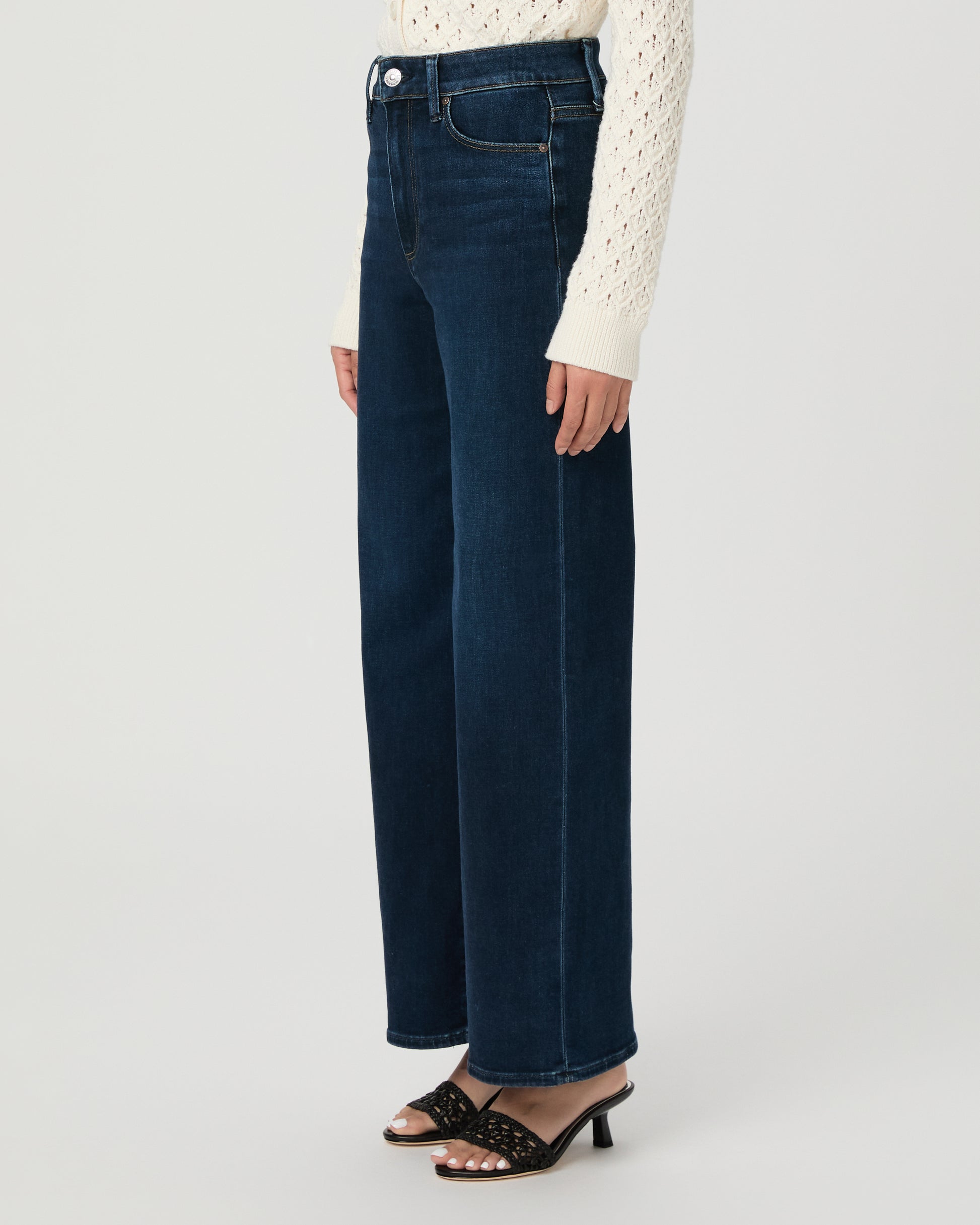 A woman modeling a cream-colored, long-sleeve open-knit cardigan paired with dark blue wide-leg jeans and black slide sandals, set against a plain light gray studio background.
