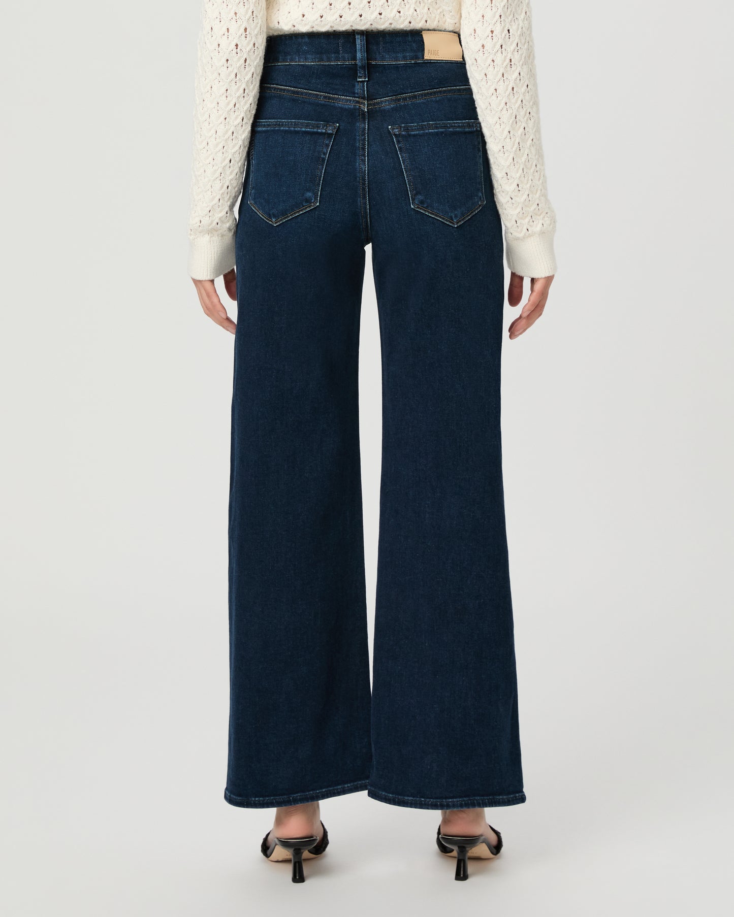 A woman modeling a cream-colored, long-sleeve open-knit cardigan paired with dark blue wide-leg jeans and black slide sandals, set against a plain light gray studio background.
