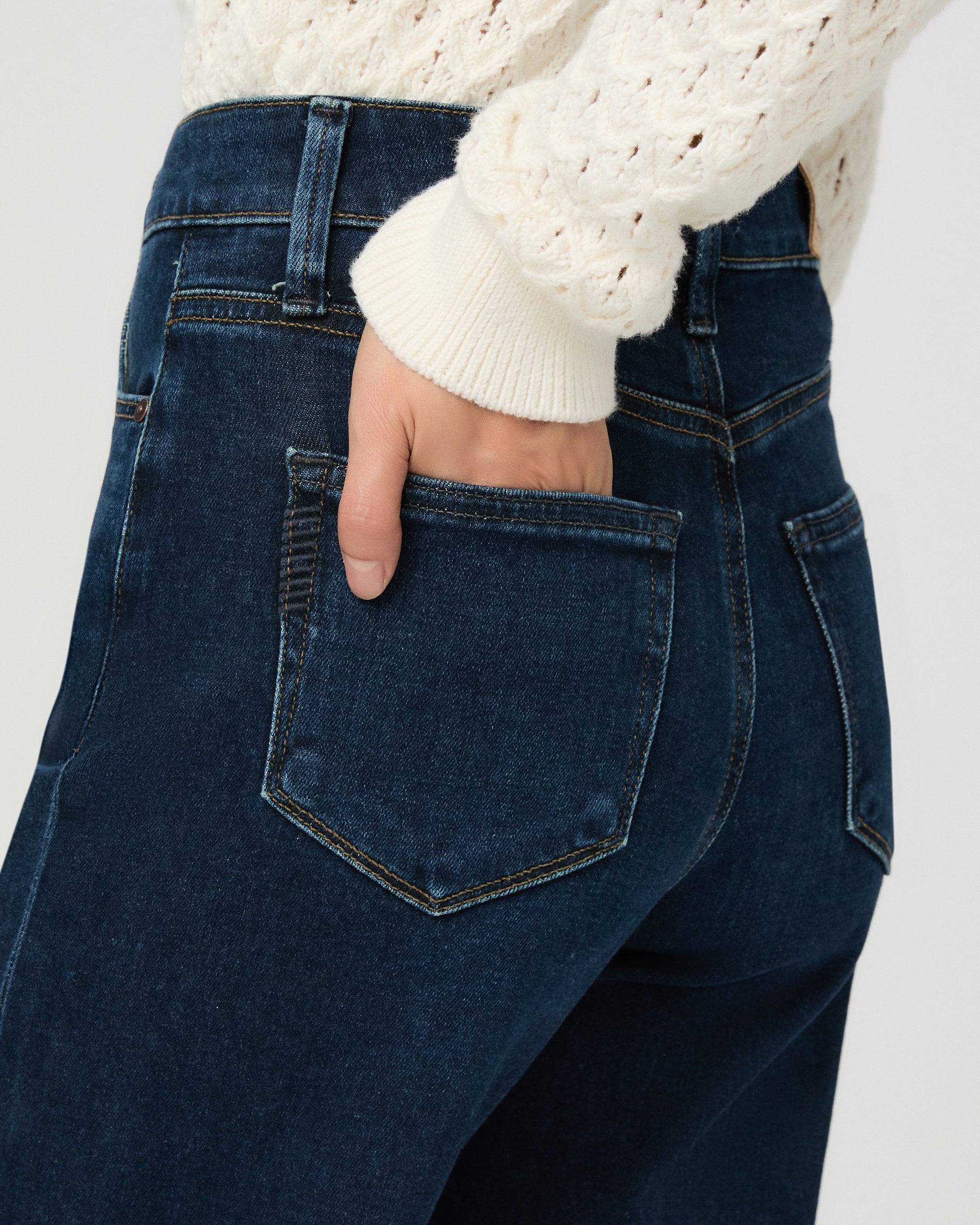 A woman modeling a cream-colored, long-sleeve open-knit cardigan paired with dark blue wide-leg jeans and black slide sandals, set against a plain light gray studio background.