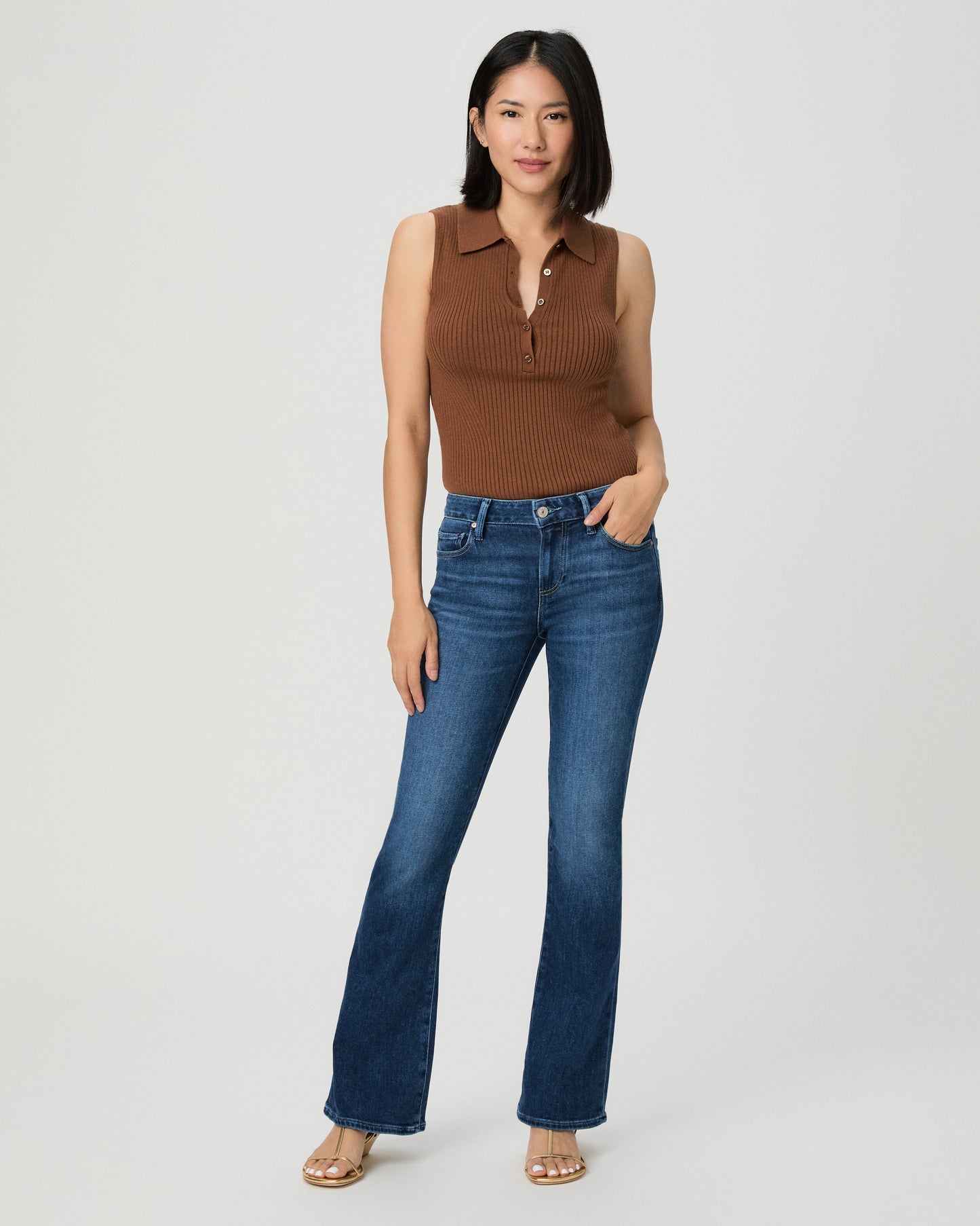 A woman modeling a brown ribbed sleeveless polo vest and dark blue flare jeans against a light gray studio background.