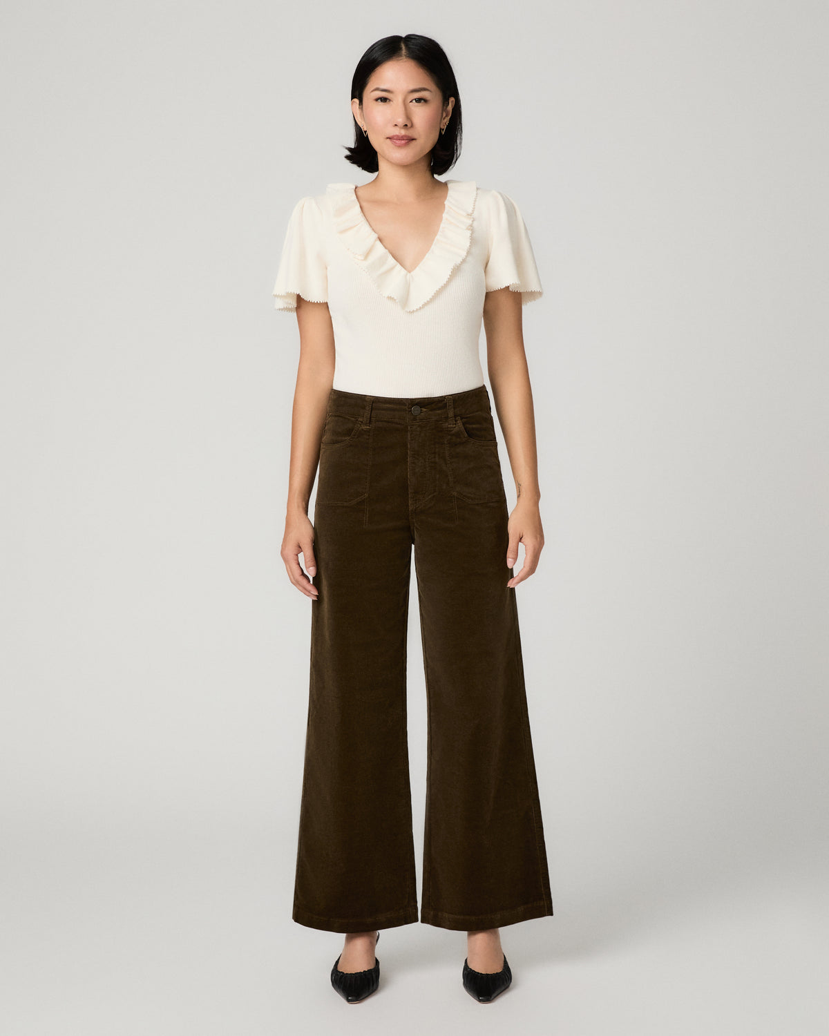A woman stands against a light gray studio background wearing a cream short-sleeve top with ruffle trim and dark brown wide-leg corduroy pants.