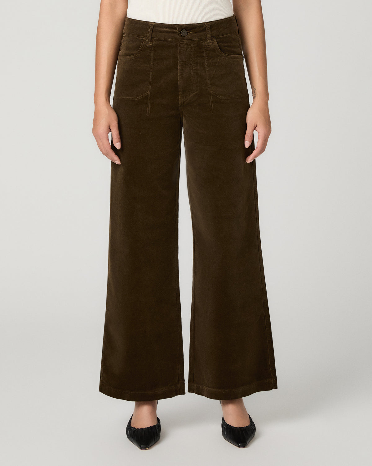 A model wearing dark brown wide-leg corduroy pants, styled with a white top and black pointed flats, against a light gray studio background.
