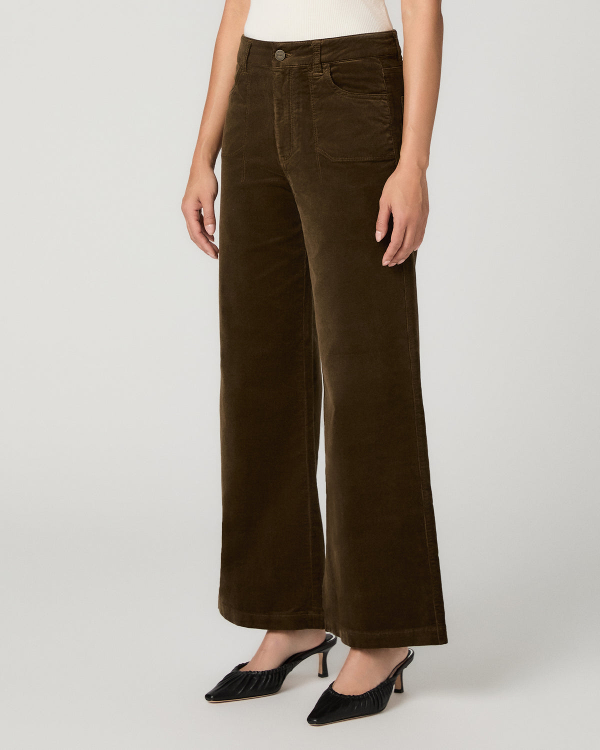 A model wearing dark brown wide-leg corduroy trousers with a white top and black pointed-toe mule heels, shown from the waist down on a light gray studio background.
