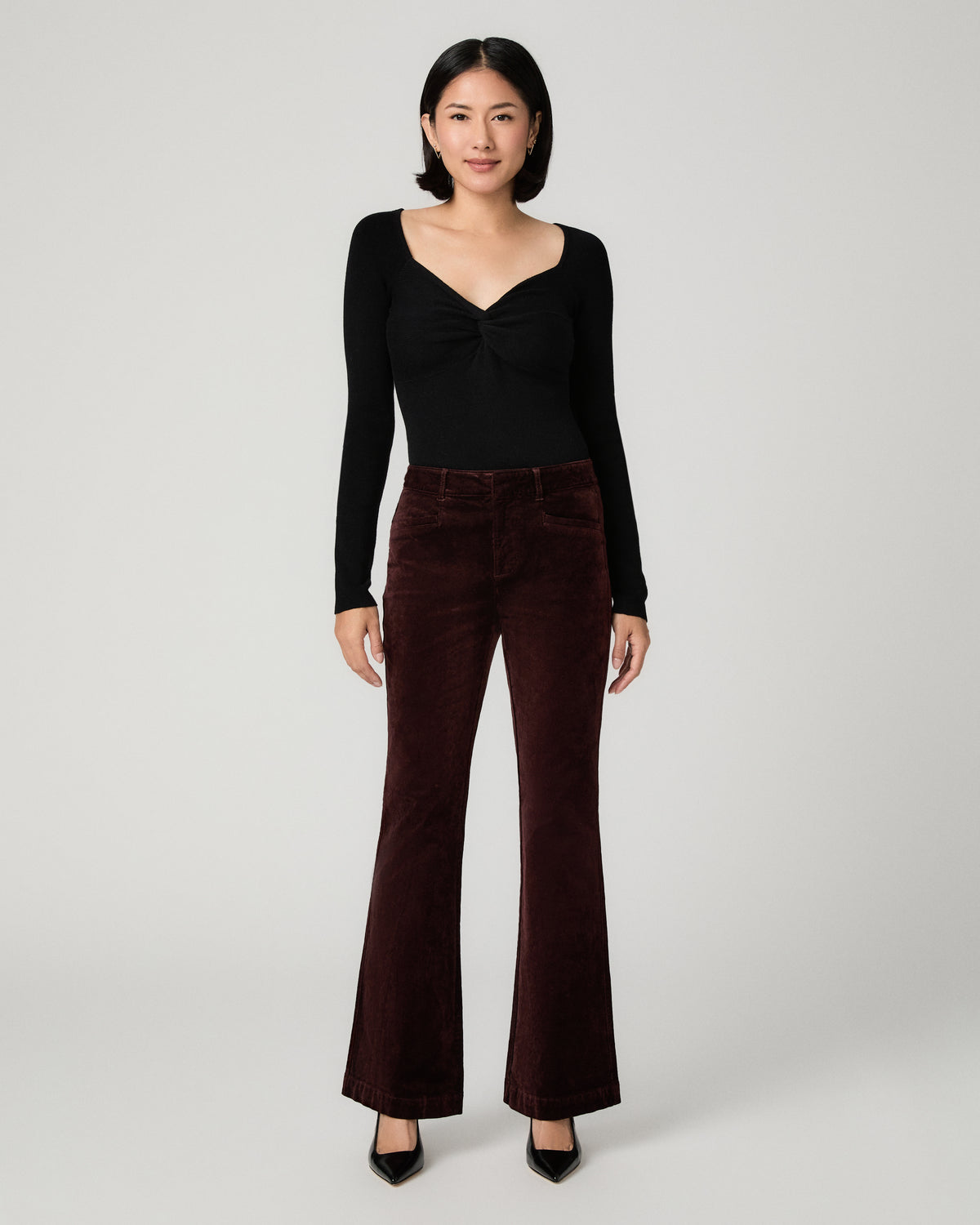 A woman stands against a light gray studio background wearing a black long-sleeve top with a gathered sweetheart neckline and dark burgundy flared pants.