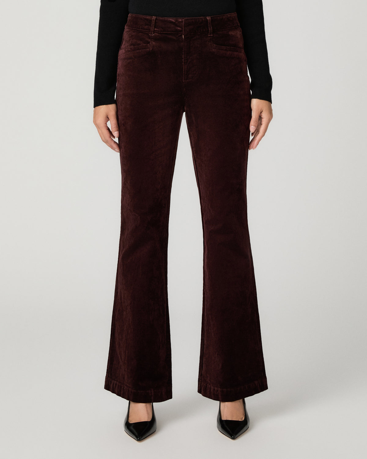 A model from the waist down wearing dark brown corduroy flare jeans, a black long-sleeve top, and black pointed-toe heels against a light gray studio background.