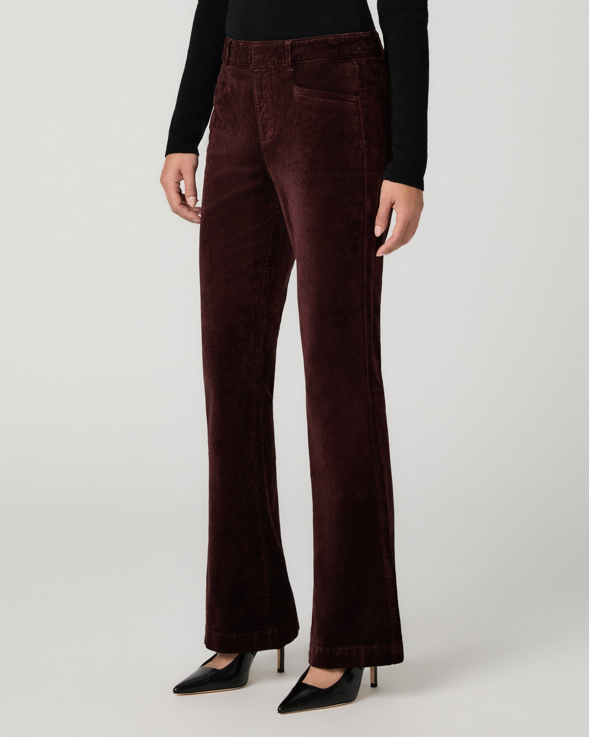 A model is shown from the waist down wearing dark burgundy velvet flare pants, a black long-sleeve top, and black pointed-toe heels against a light gray studio background.
