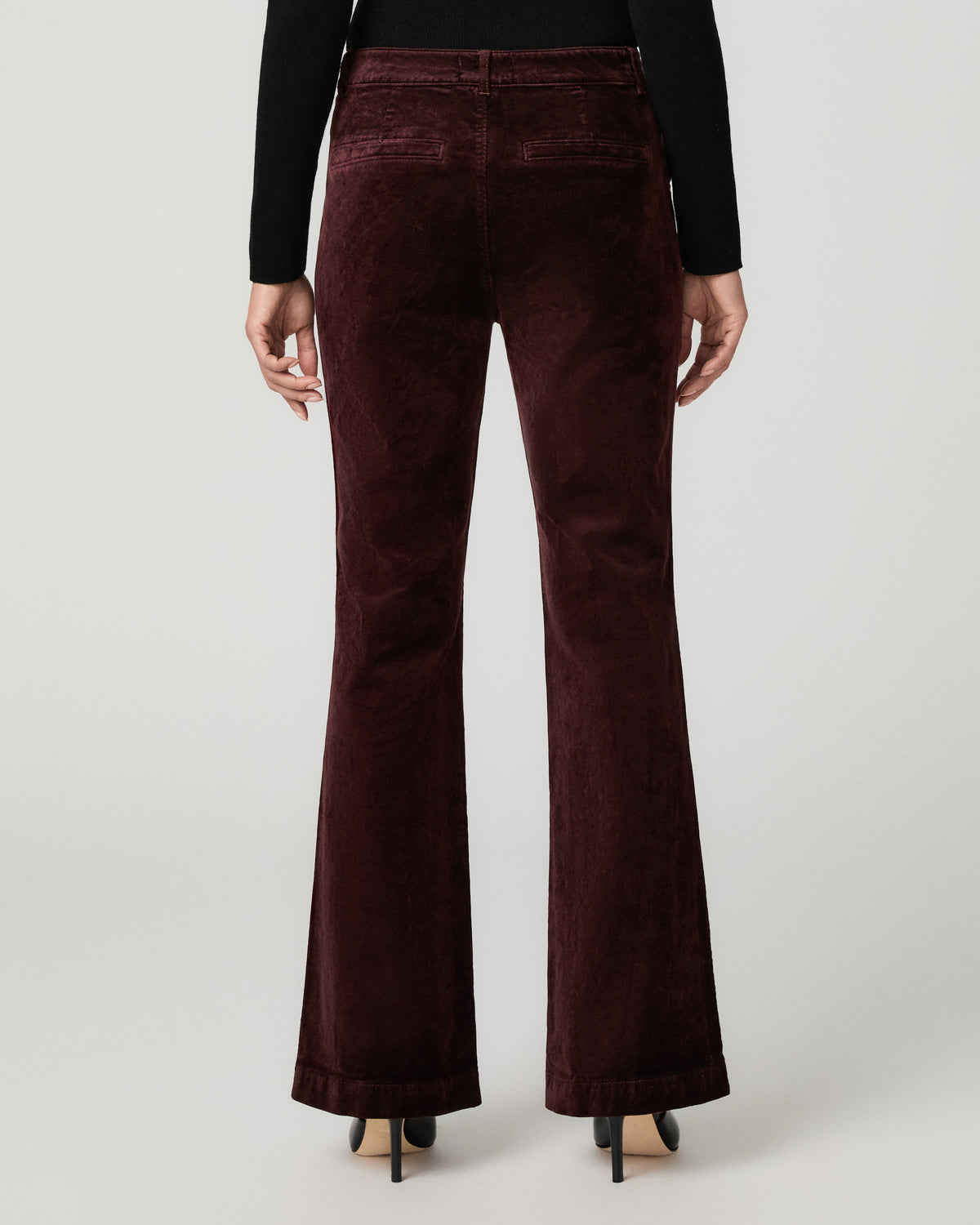 A person shown from the back wearing high-waisted, wide-leg flared trousers in a deep burgundy velvet fabric, paired with a black long-sleeve top and black heels, against a light gray studio background.