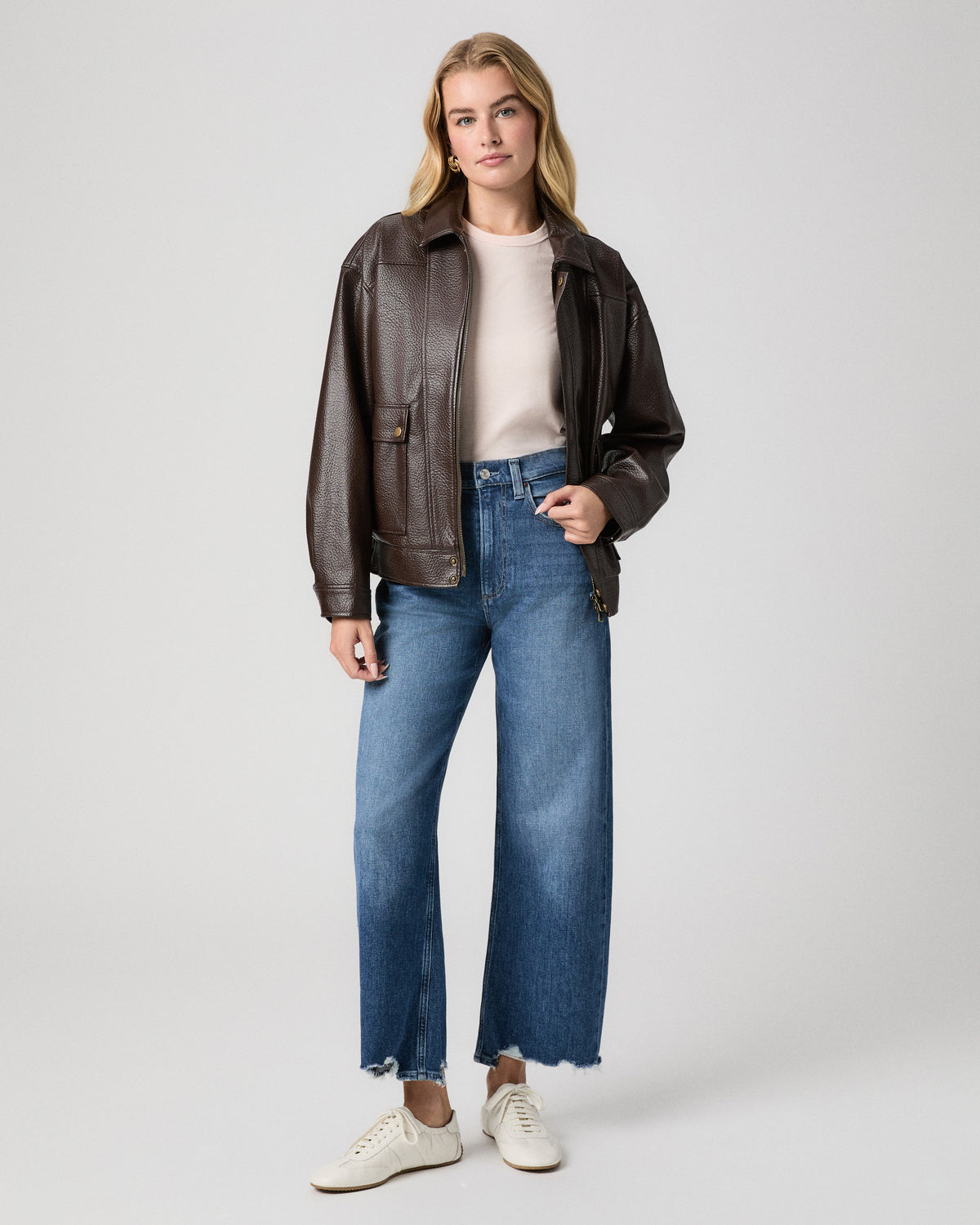 Arellia Ankle Barrel Leg Jean in Stoneview medium wash with frayed hem; model stands with a hand in her pocket, in a brown leather jacket, cream tee and white sneakers