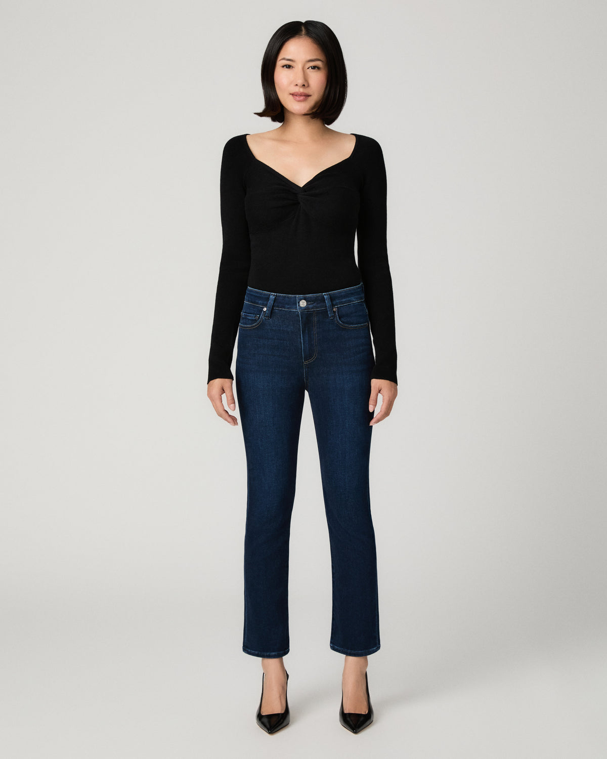 A model wearing a black long-sleeve top with a twisted front detail and dark blue straight-leg jeans, standing against a light gray studio background.