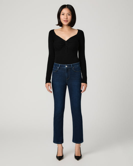 A model wearing a black long-sleeve top with a twisted front detail and dark blue straight-leg jeans, standing against a light gray studio background.