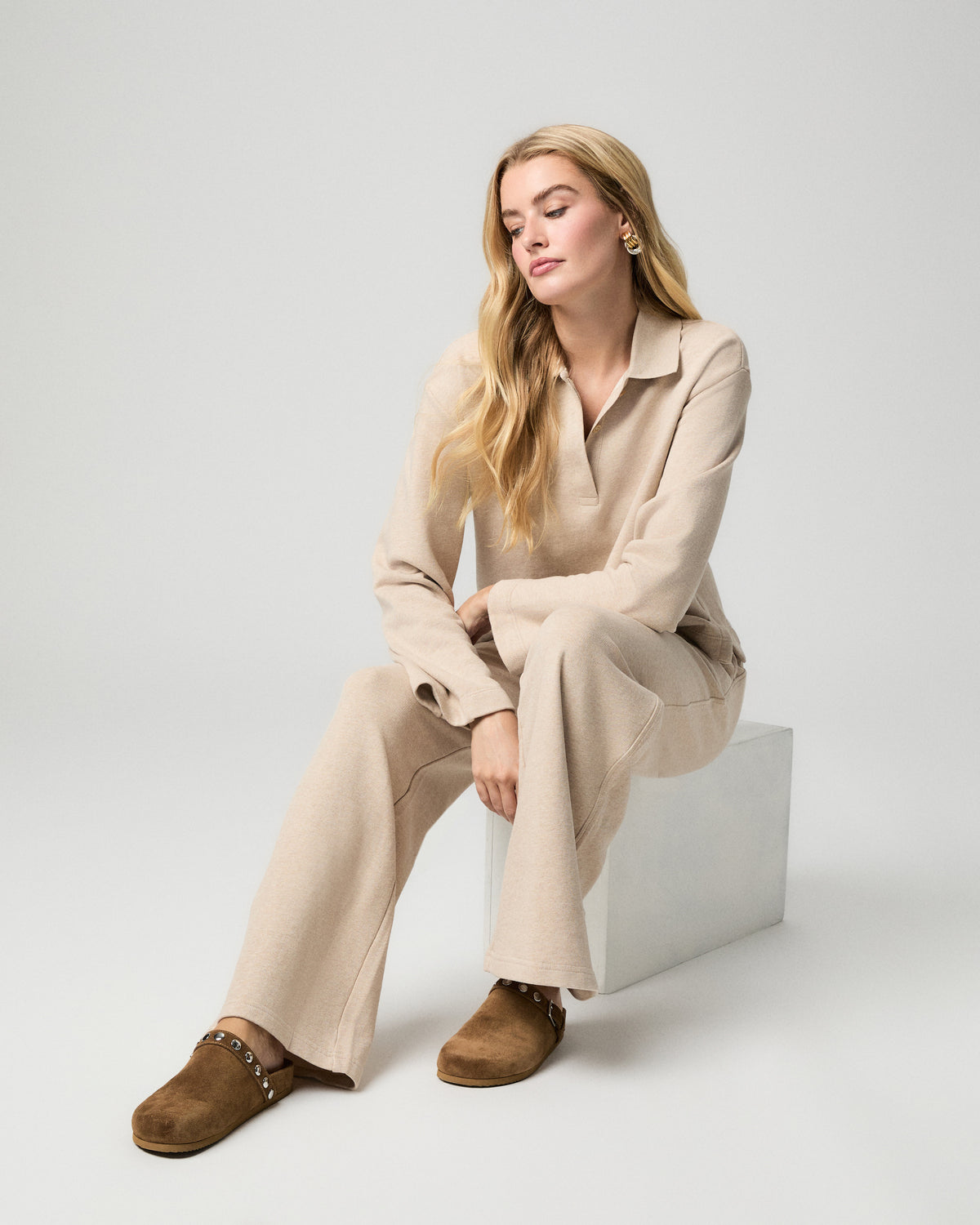 Model seated on a white block, wearing the Whistler Pant in Heathered Oatmeal with a matching collared knit top and brown suede clogs