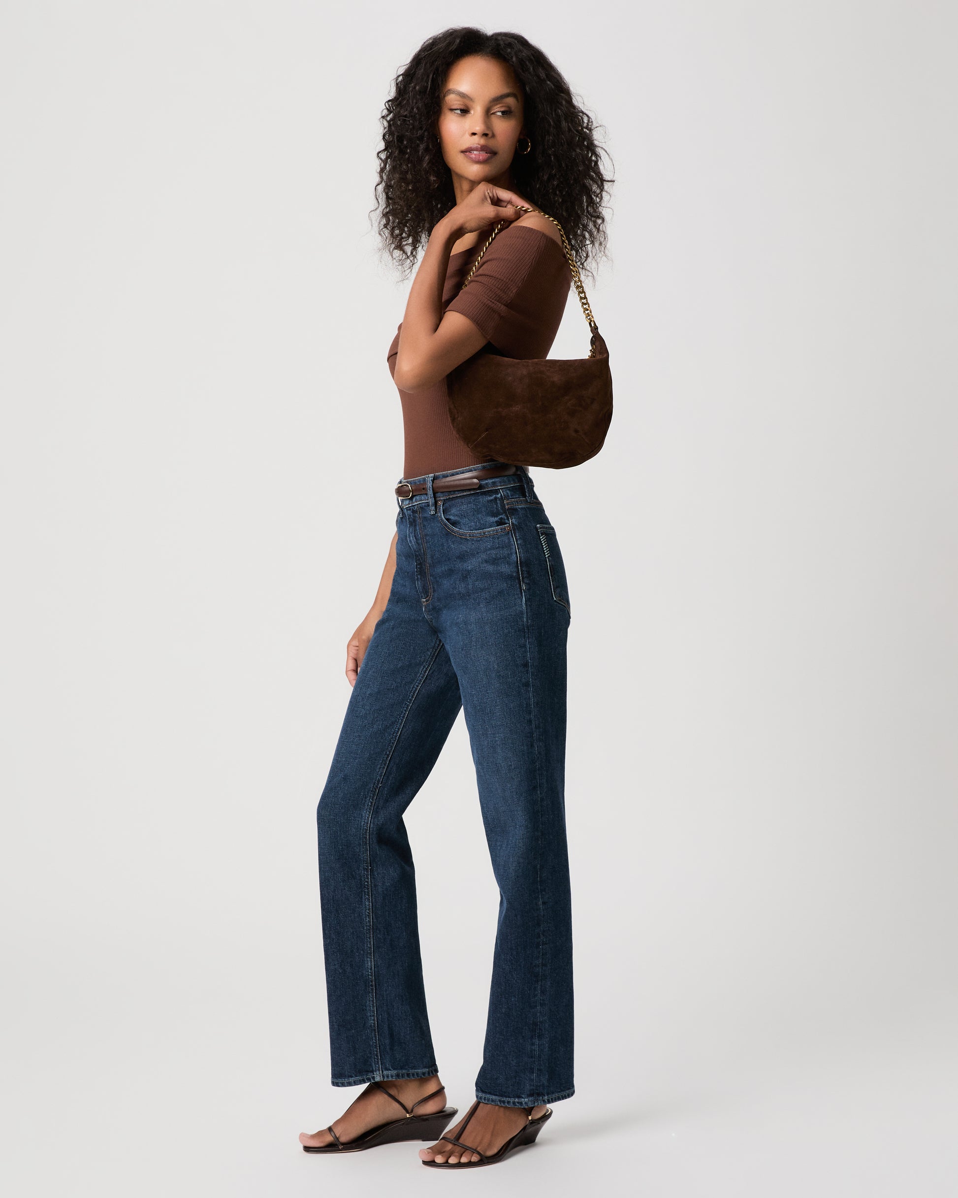 Women wearing brown top paired with blue jeans holding a brown green bag against a white background 