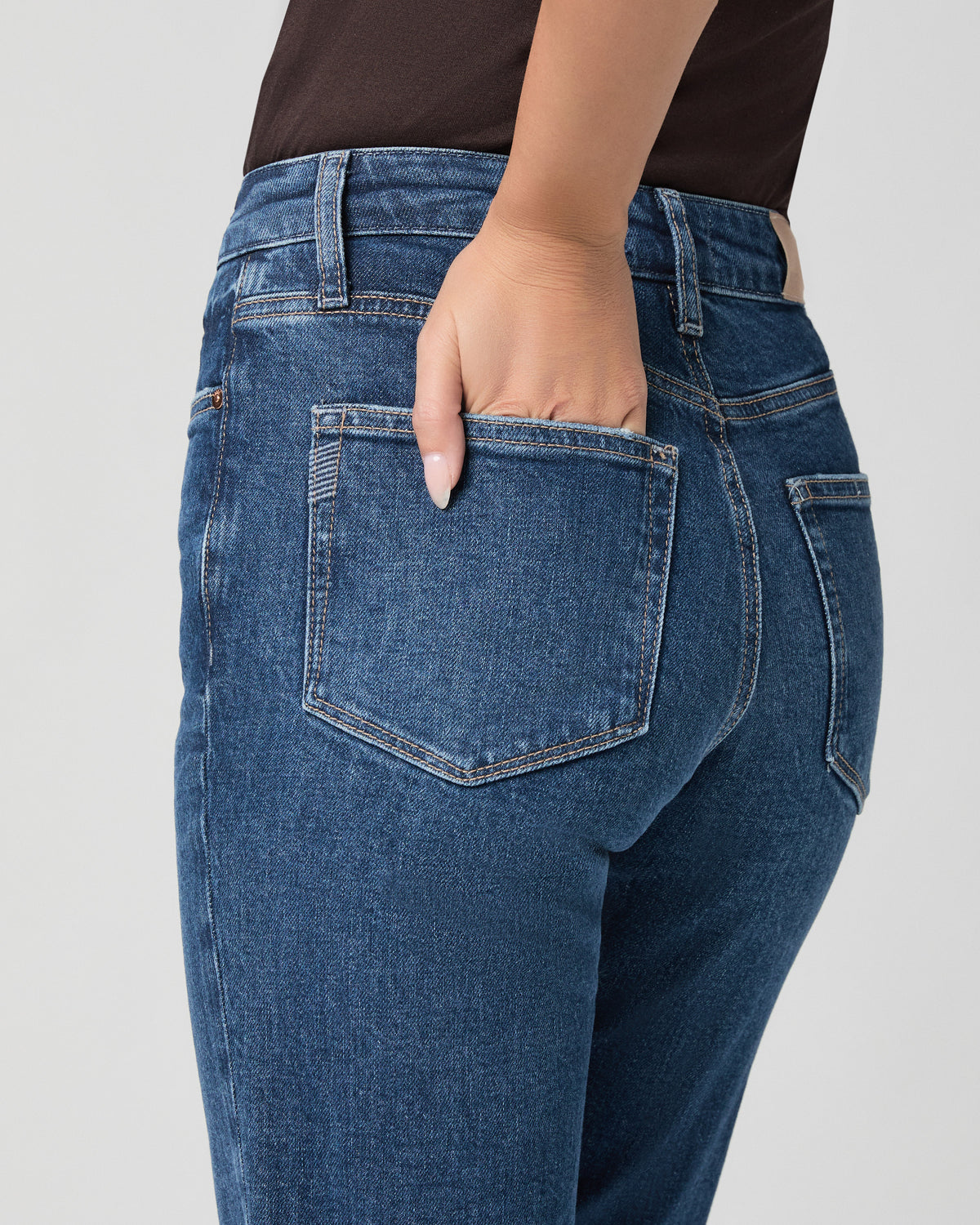 Close-up of the back pocket of the Stevie Straight Jean in Soundcheck dark wash; model has one hand in the pocket and wears a brown top