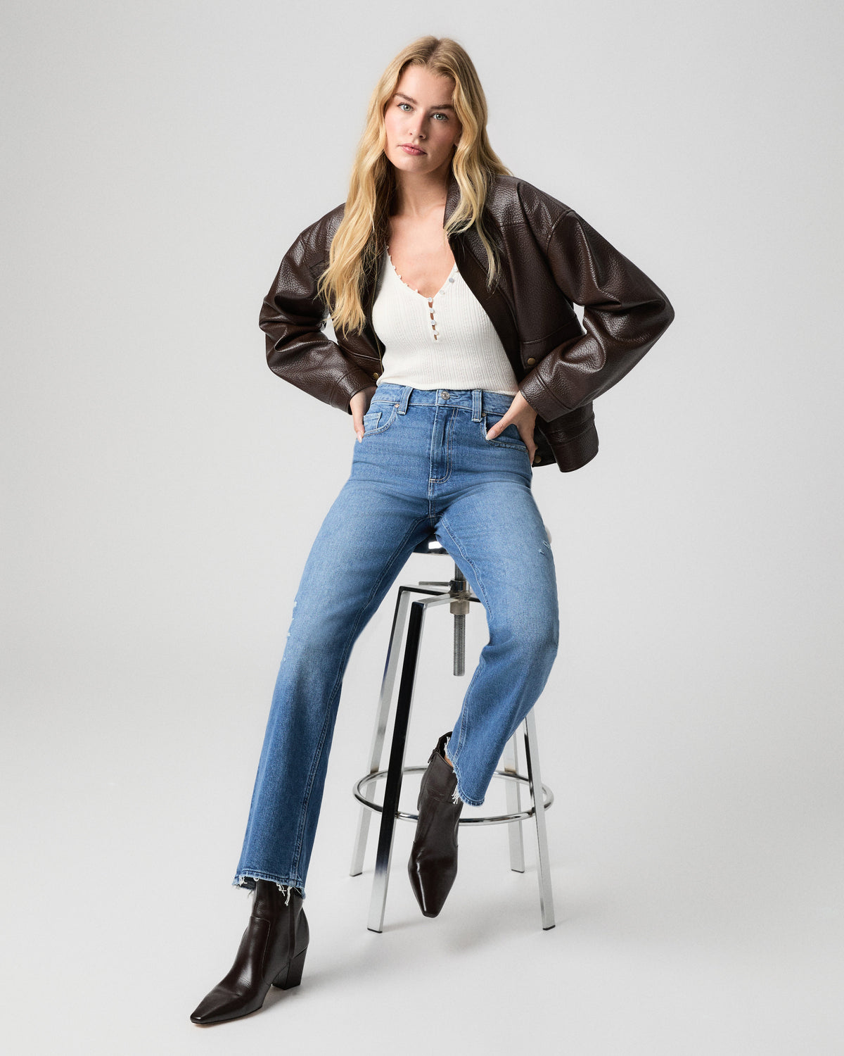 Stevie Ankle Straight Jean in Frittata Dest light-medium wash; model sits on a stool in a brown leather jacket, white henley and black ankle boots