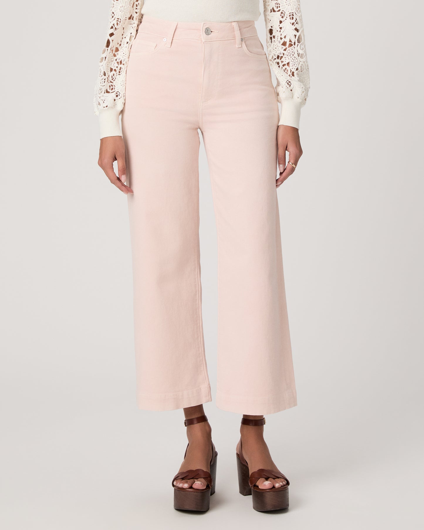 Woman wearing a white lace top, pink pants, and brown platform sandals on a light gray background