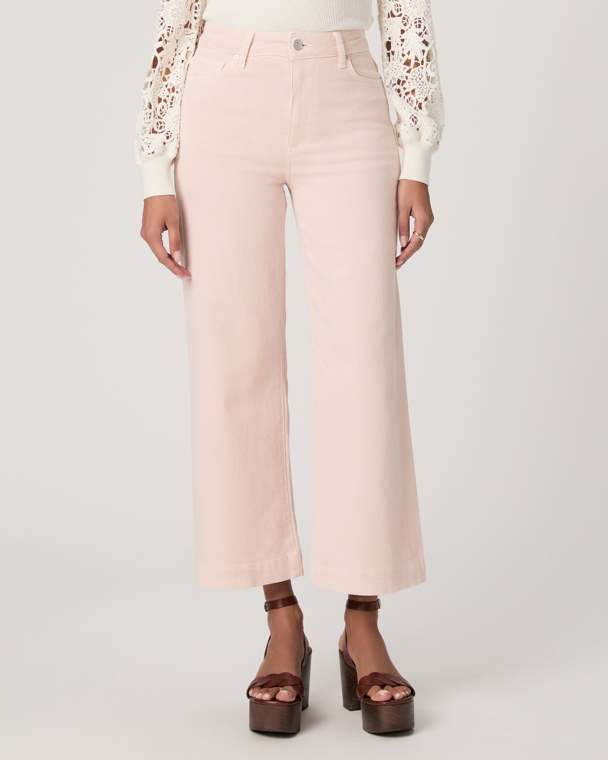 Woman wearing a white lace top, pink pants, and brown platform sandals on a light gray background