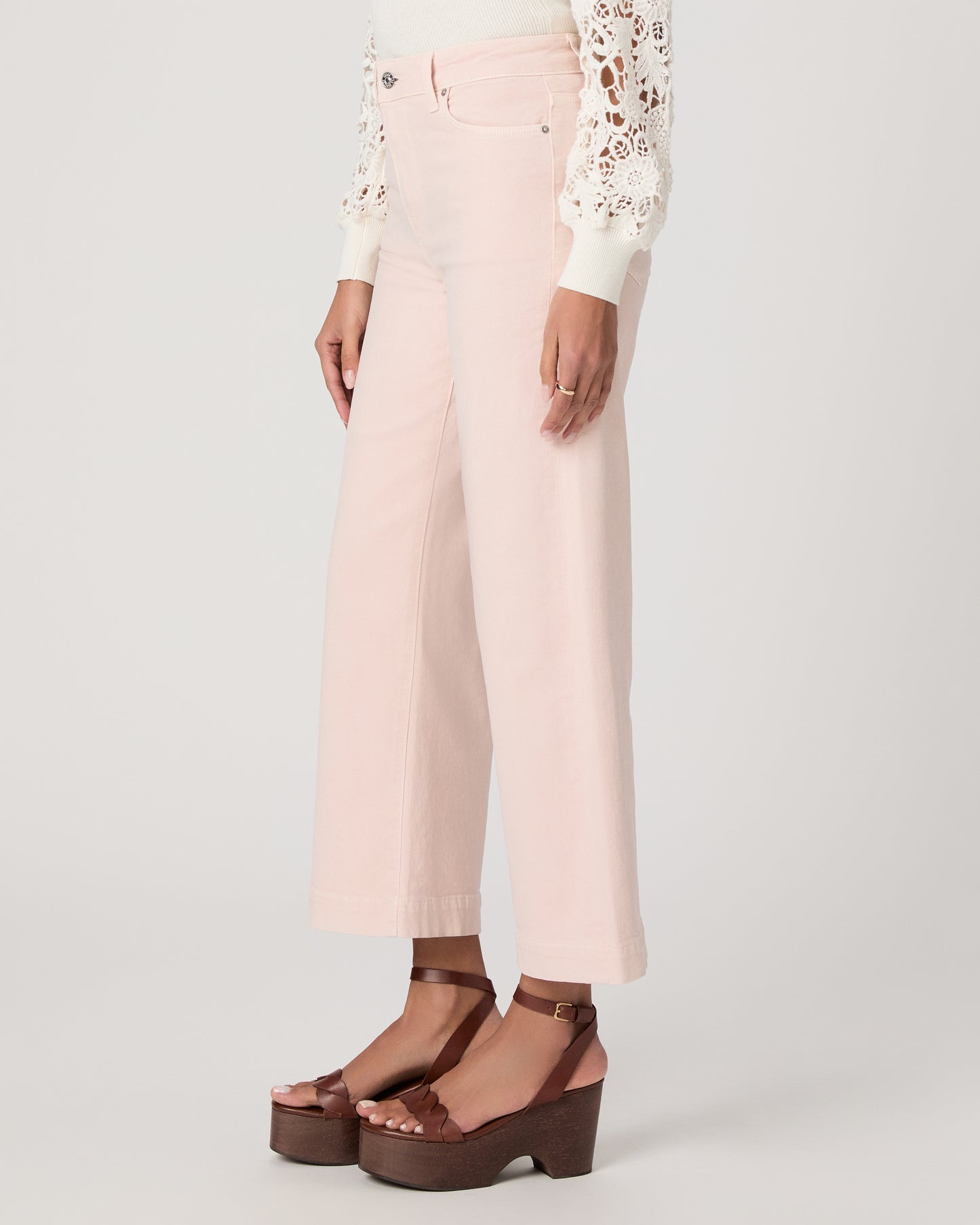 Woman wearing a white lace top, pink pants, and brown platform sandals on a light gray background