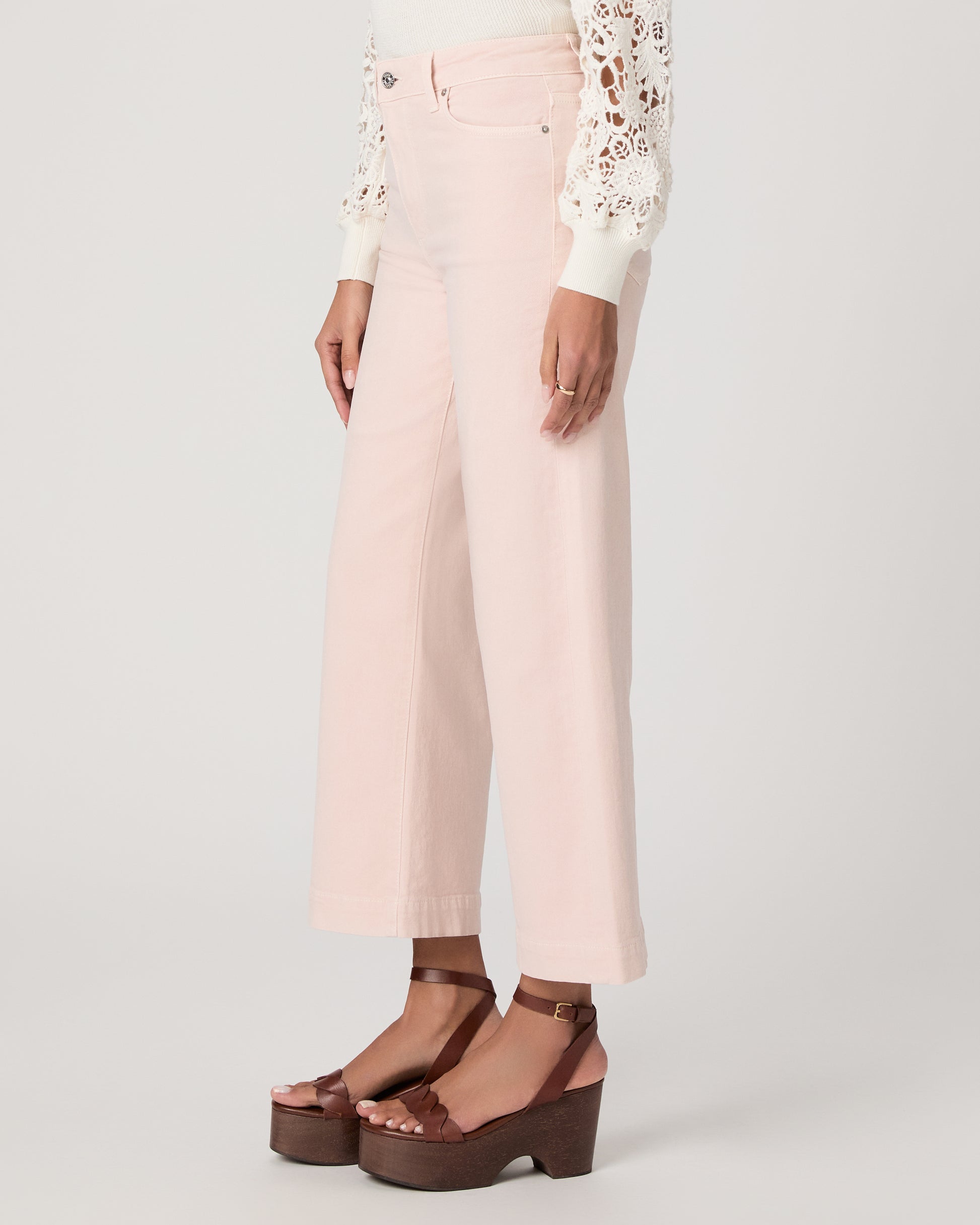 Woman wearing a white lace top, pink pants, and brown platform sandals on a light gray background