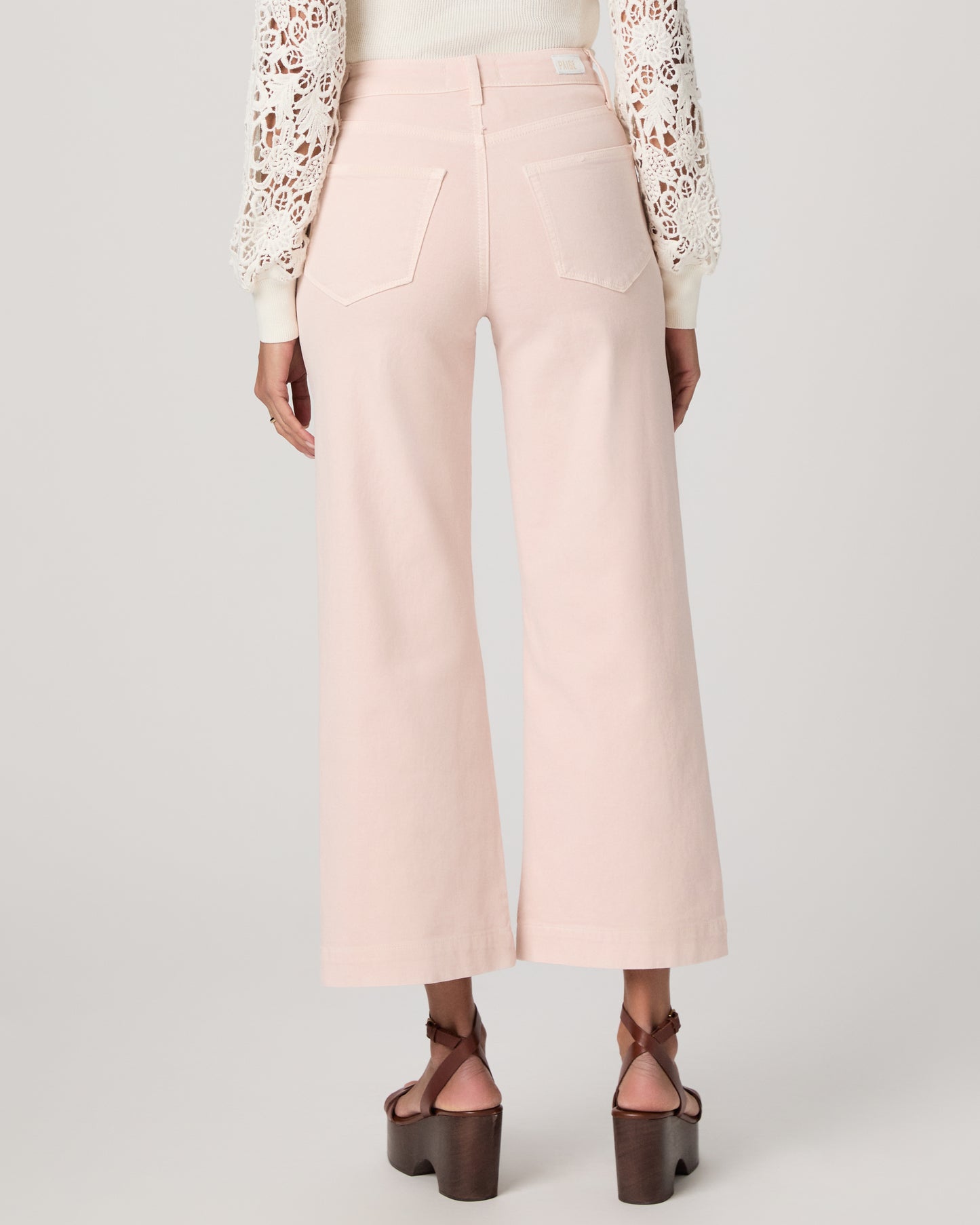 Woman wearing a white lace top, pink pants, and brown platform sandals on a light gray background
