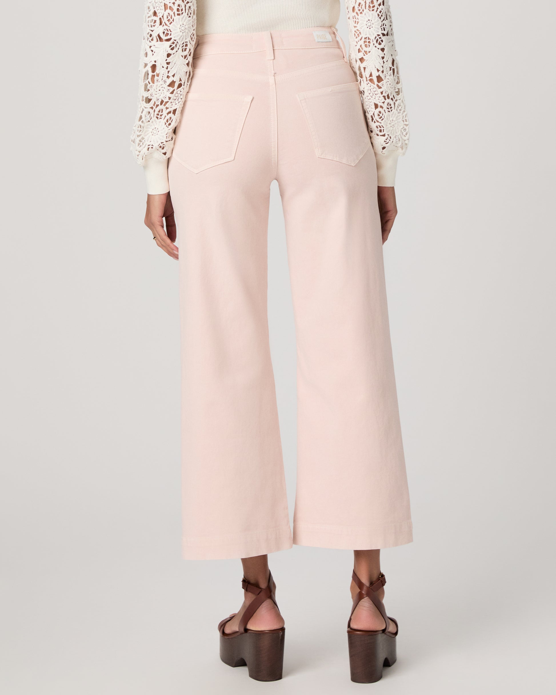 Woman wearing a white lace top, pink pants, and brown platform sandals on a light gray background