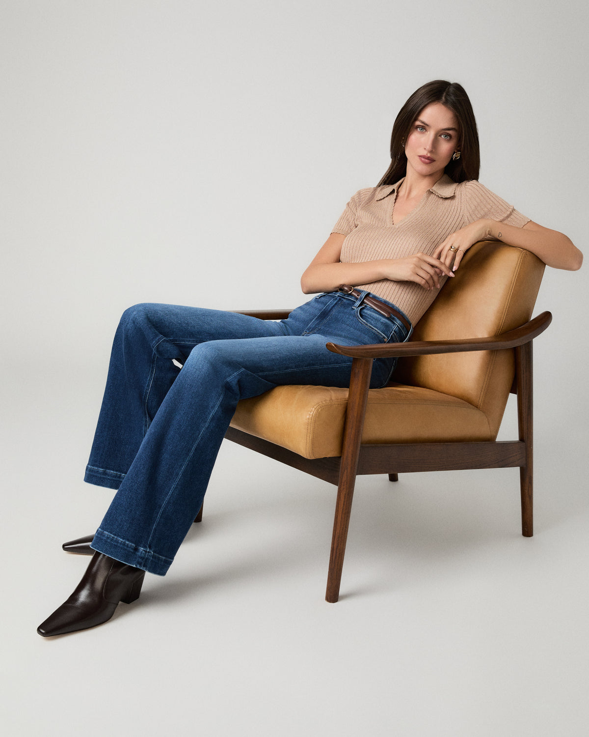 A woman sits in a mid-century modern armchair wearing a beige ribbed short-sleeve polo shirt, blue flare jeans, and dark brown pointed-toe boots against a light gray studio background.