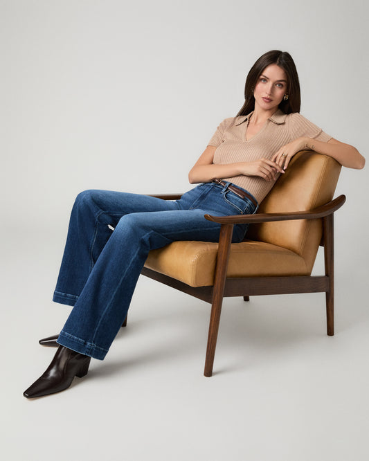 A woman sits in a mid-century modern armchair wearing a beige ribbed short-sleeve polo shirt, blue flare jeans, and dark brown pointed-toe boots against a light gray studio background.