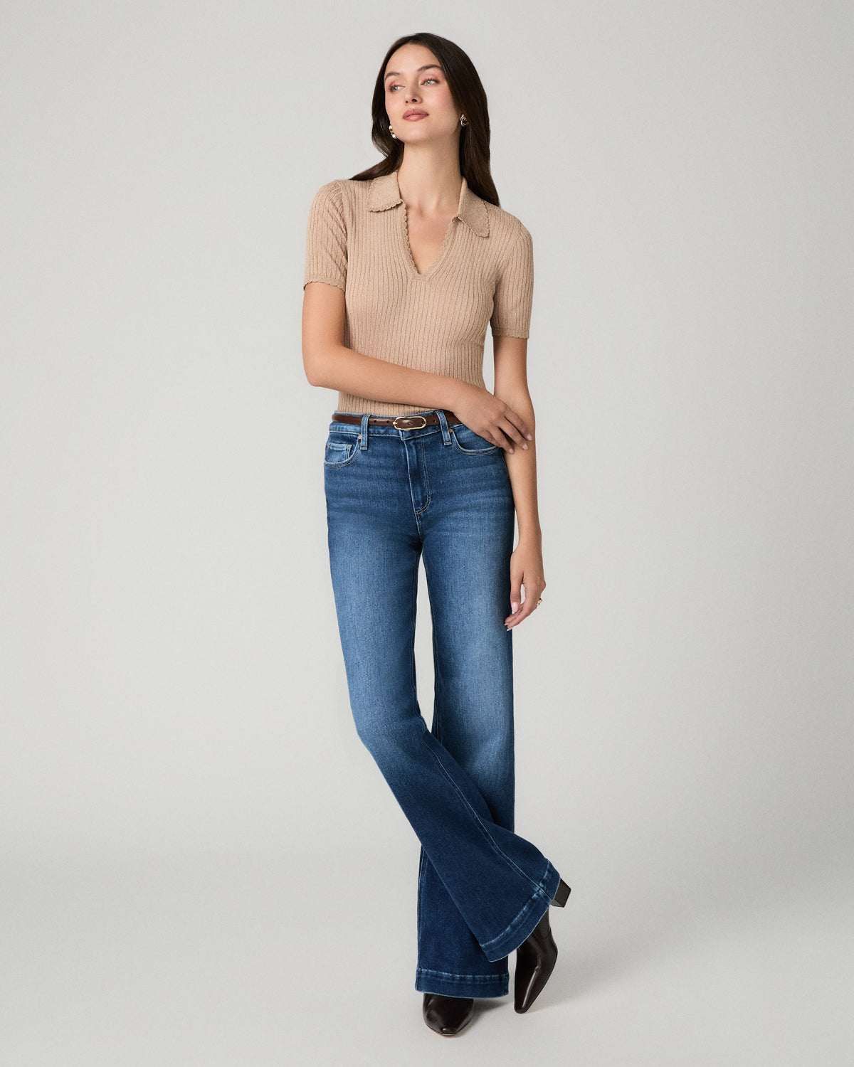 A woman modeling a beige short-sleeve ribbed knit polo shirt and dark blue flare jeans against a light gray studio background.