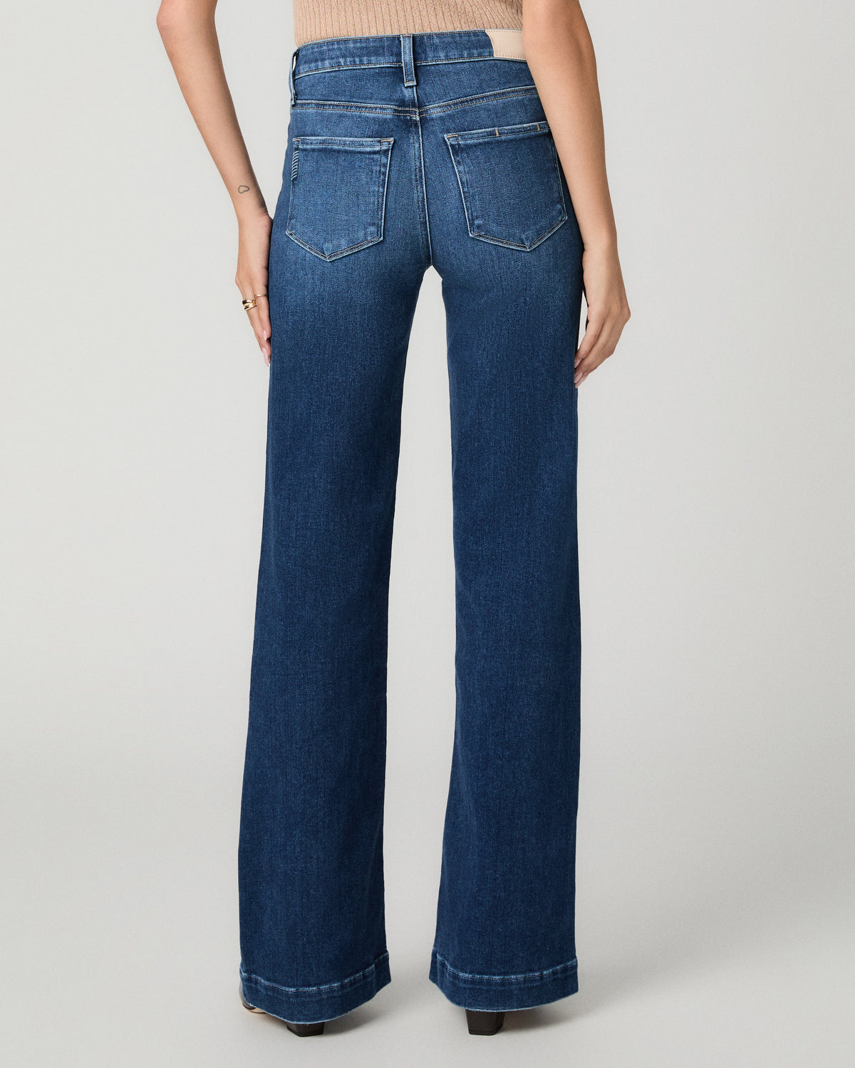 A model shown from the back wearing dark blue wide-leg denim jeans, paired with a beige ribbed top and black heels, against a light gray studio background.