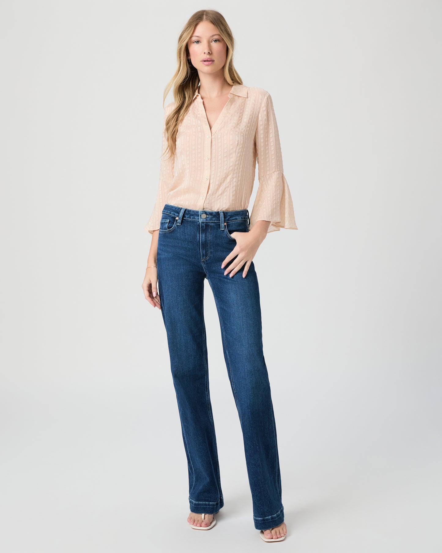 Woman wearing a beige blouse and blue jeans paired with white sandals on a white background