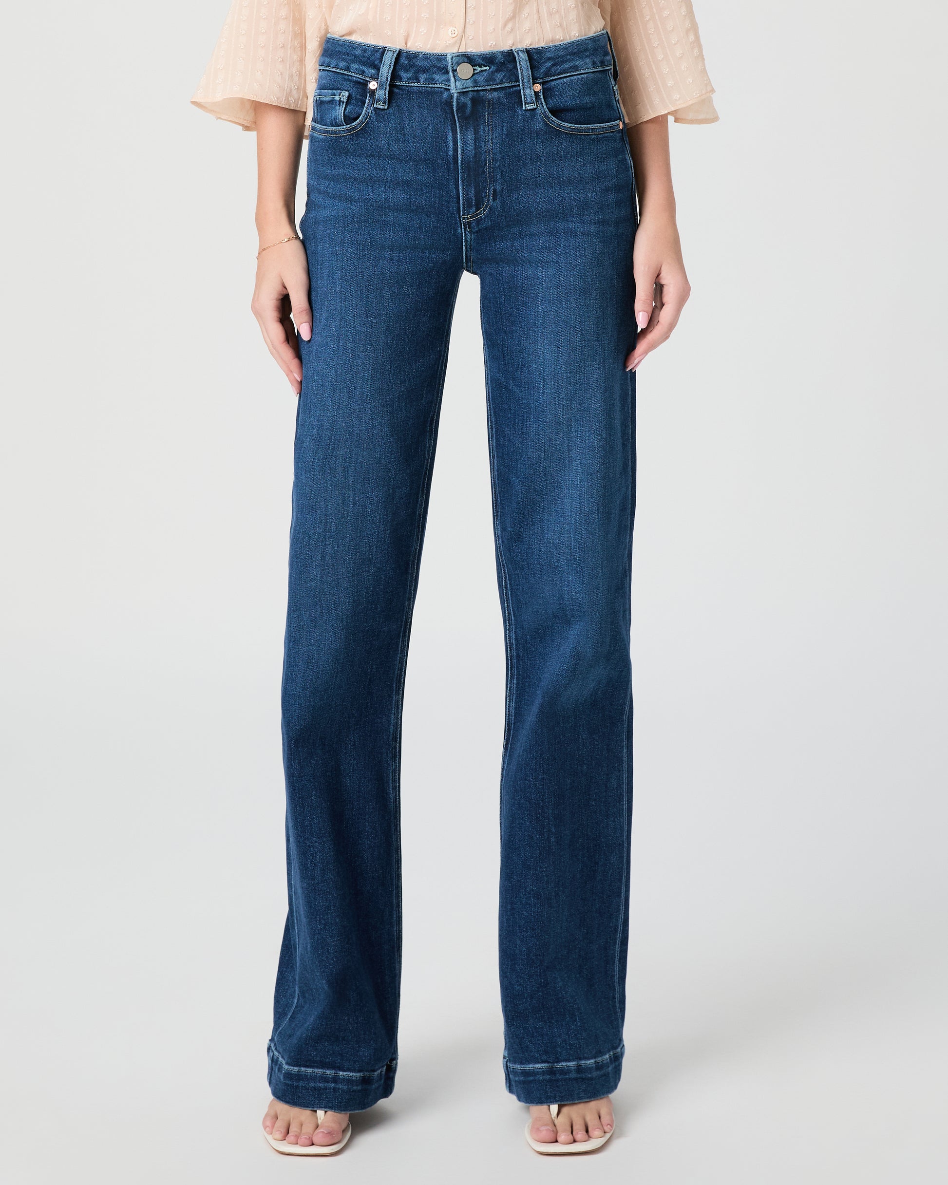 Woman wearing a beige blouse and blue jeans paired with white sandals on a white background